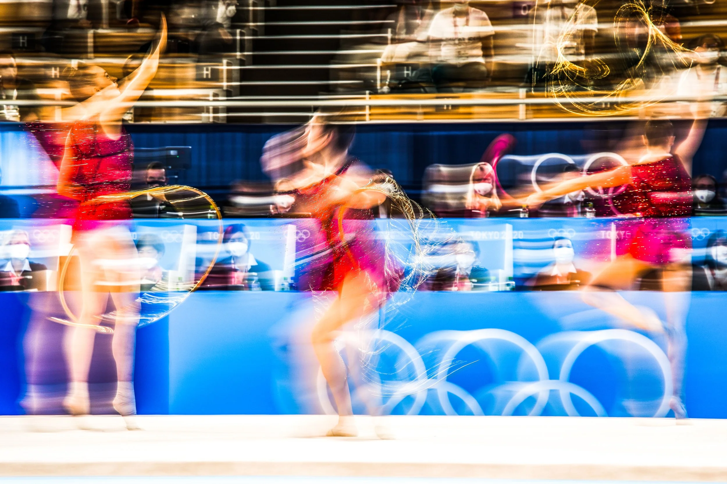 Three rhythmic gymnasts performing with hoops during a competition at the Tokyo 2020 Olympics, with blurred motion and spectators in the background.