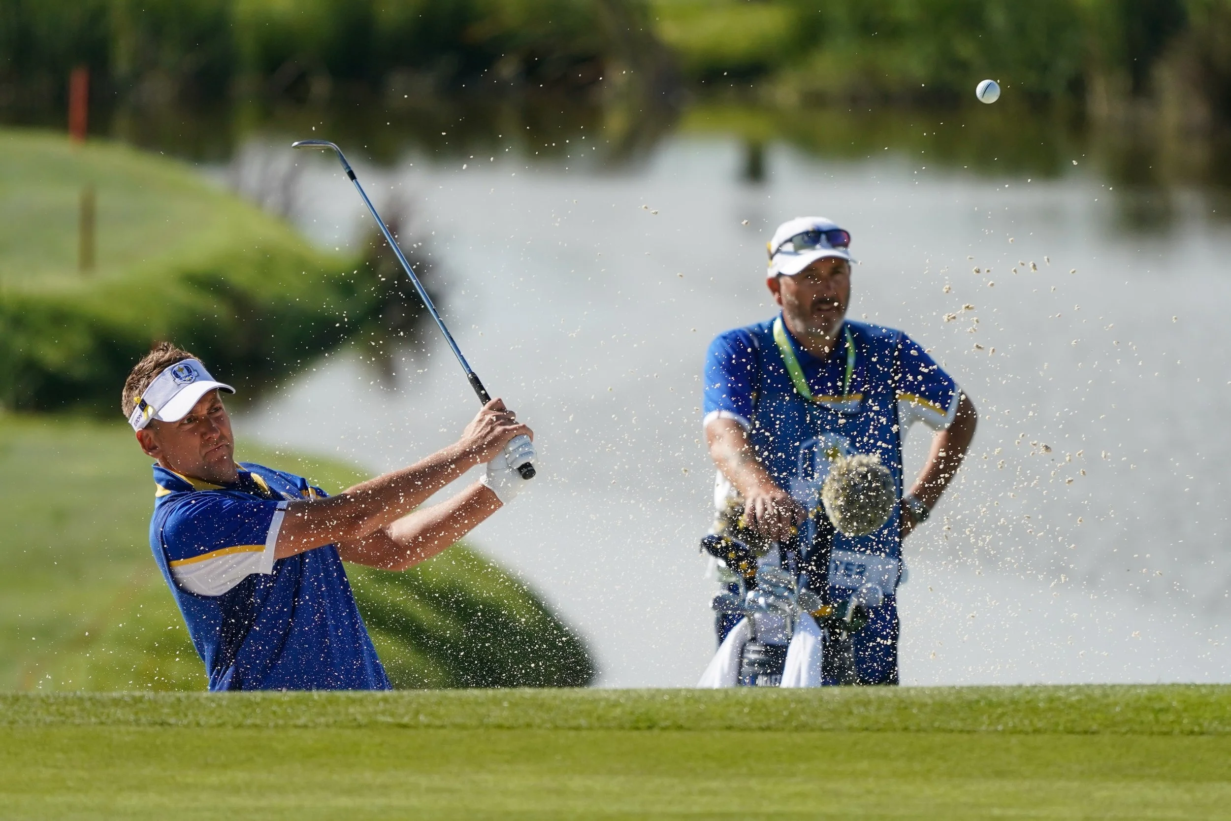 Two men in blue shirts playing golf near a water hazard, with one striking the ball and the other watching