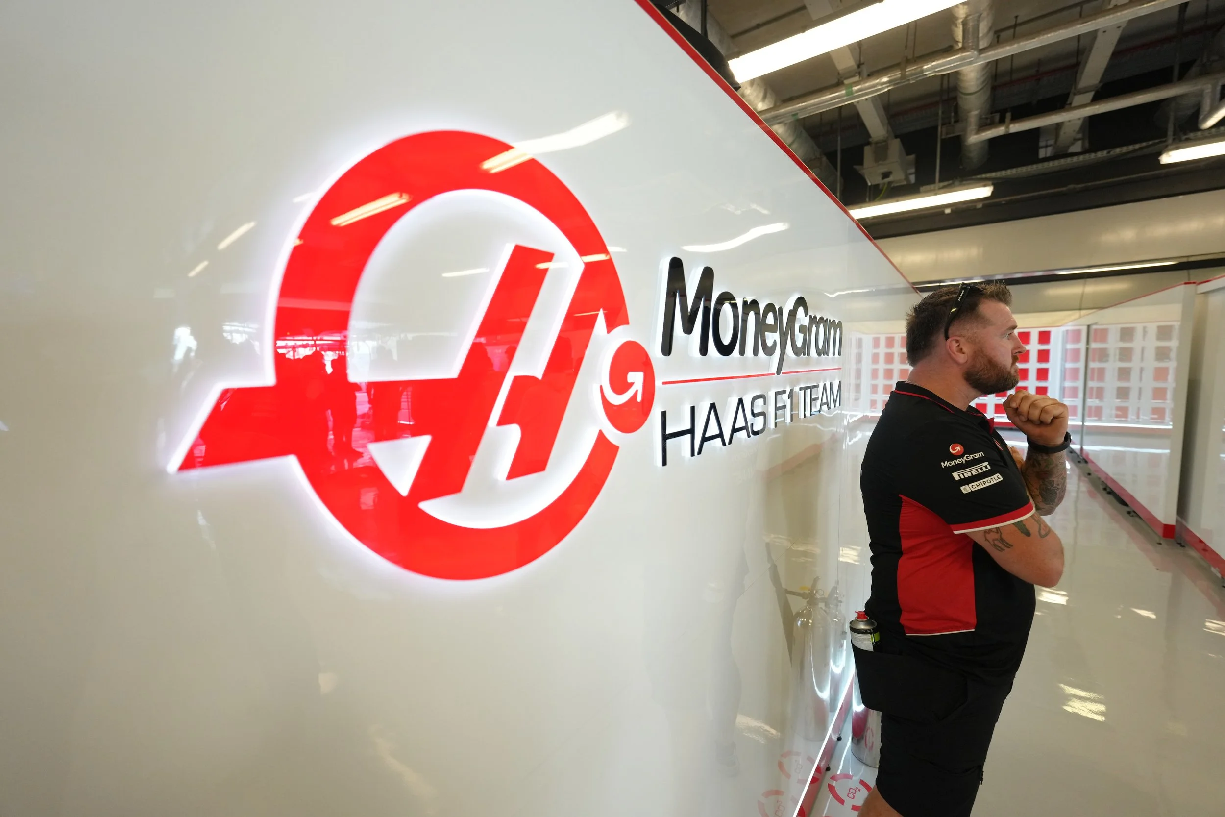 A person standing next to a large white wall with the MoneyGram logo and name, wearing a black and red shirt, in an indoor racing or garage setting.