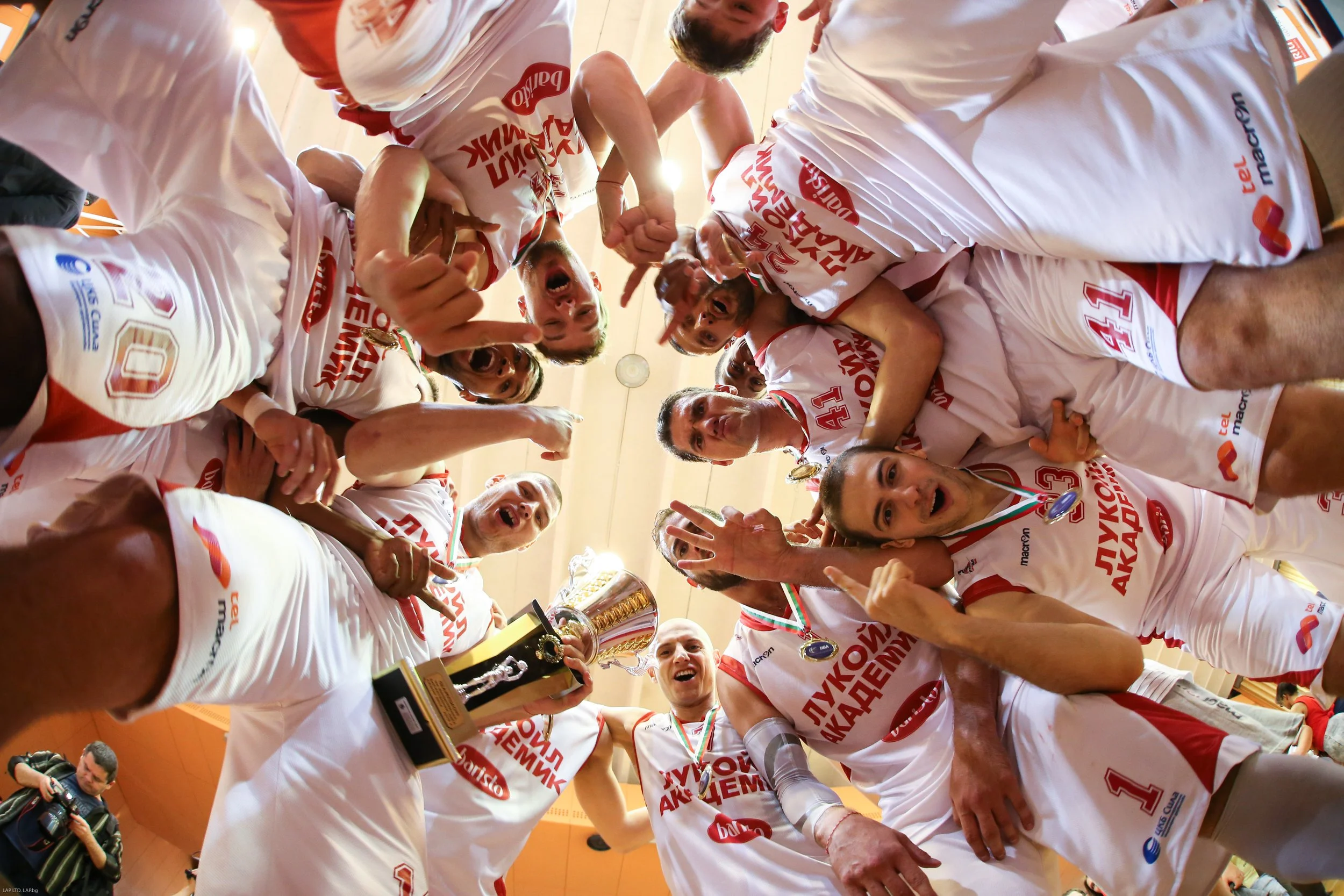 A group of athletes in white and red uniforms celebrating in a circle, one holding a trophy, some wearing medals, inside a sports hall.