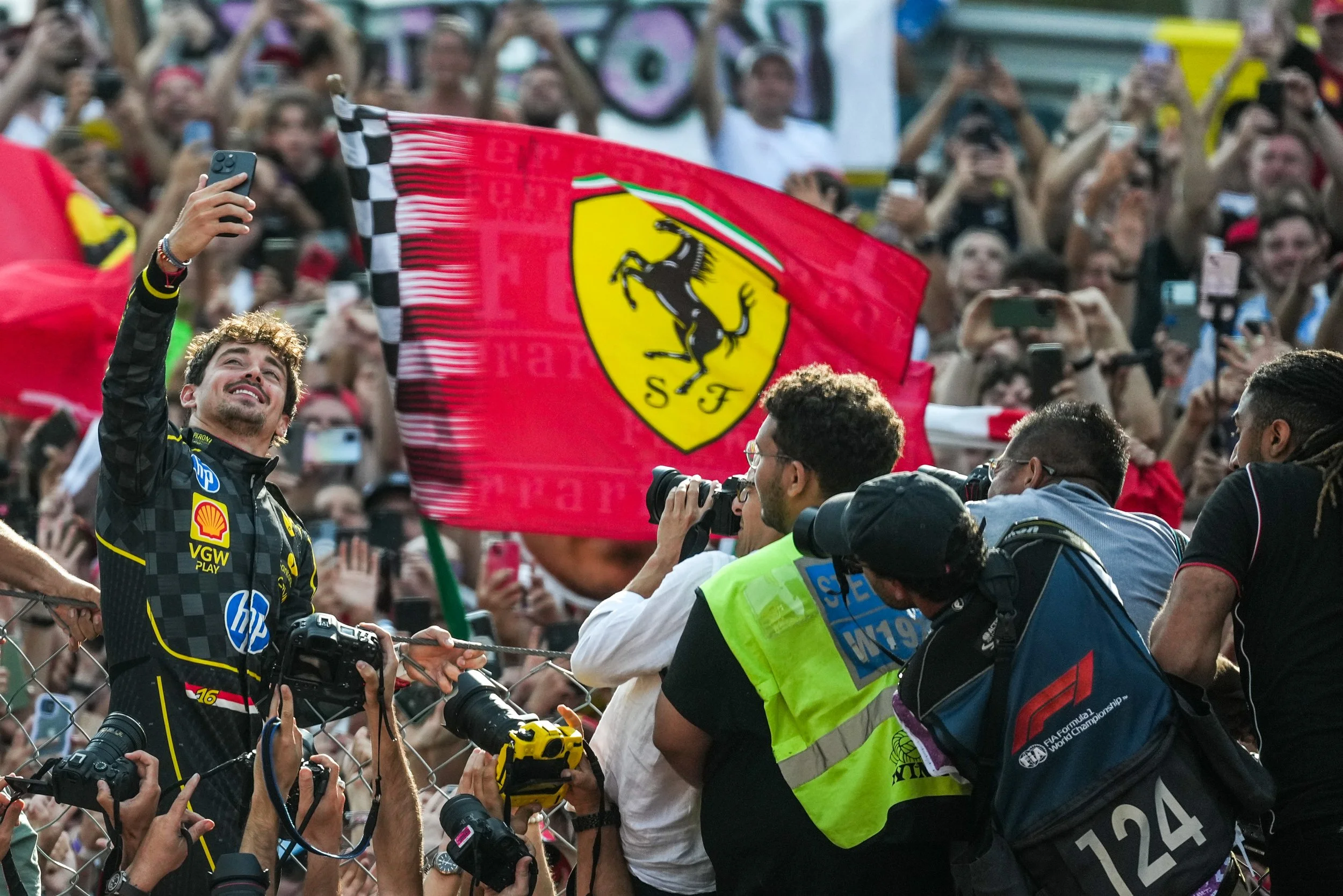 A man in a racing suit taking a selfie in front of a large Ferrari flag at a crowded event.
