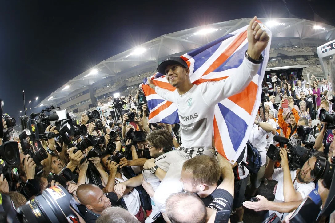 A race car driver celebrating on a podium, holding a Union Jack flag, surrounded by photographers and spectators at night.