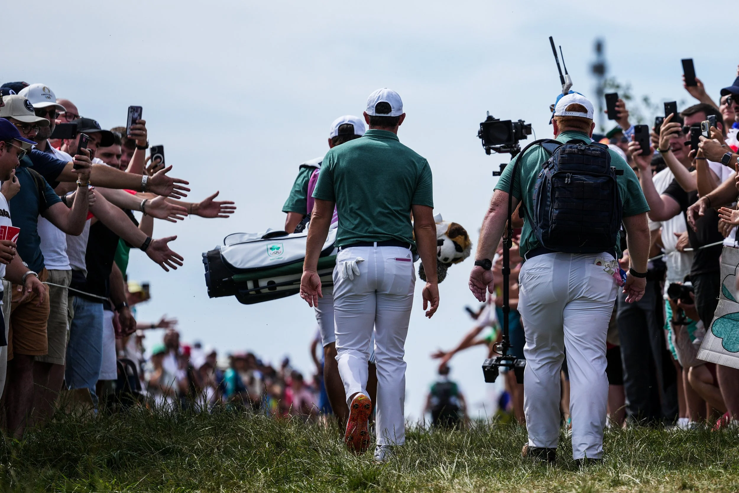 Professional golfers walking onto the fairway with their caddies, surrounded by numerous spectators taking photos and reaching out.