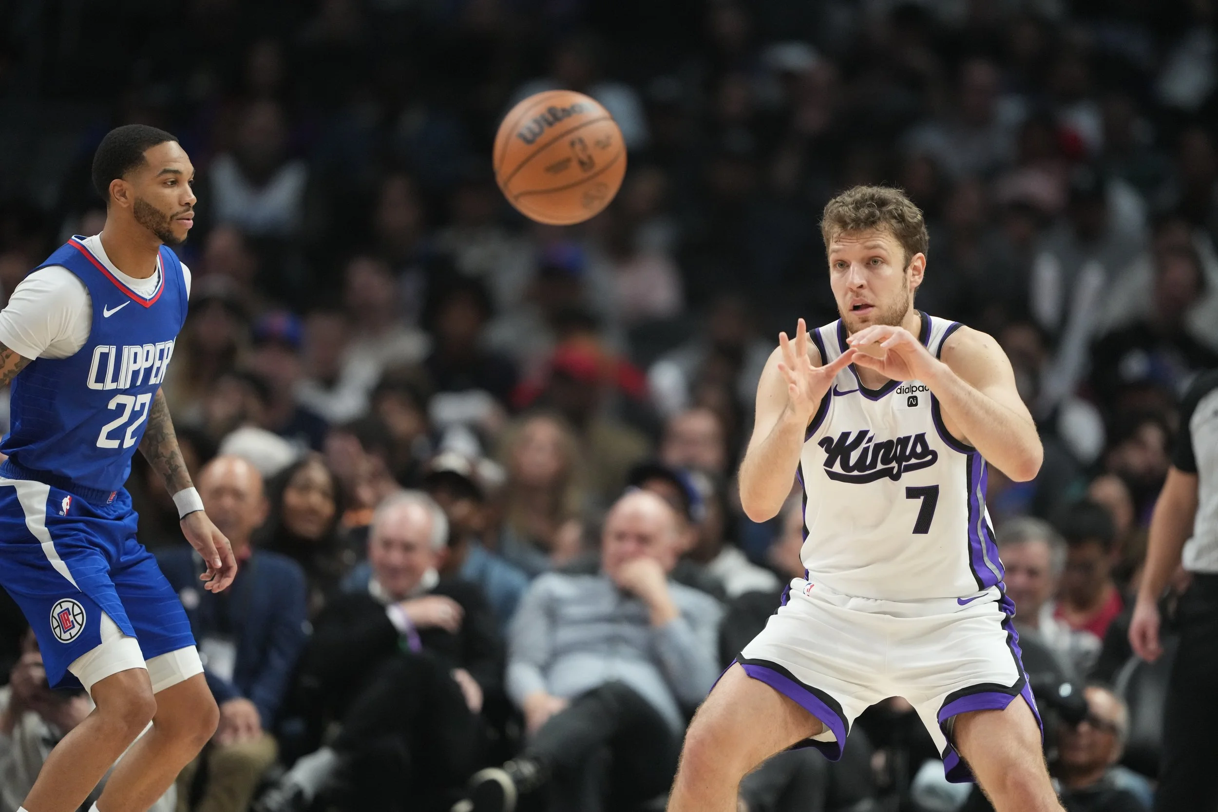 Two basketball players on the court, with one wearing a white jersey and the other in a blue jersey, during a game. The player in white is preparing to catch a basketball in mid-air, while the player in blue watches. A crowd is visible in the backgro