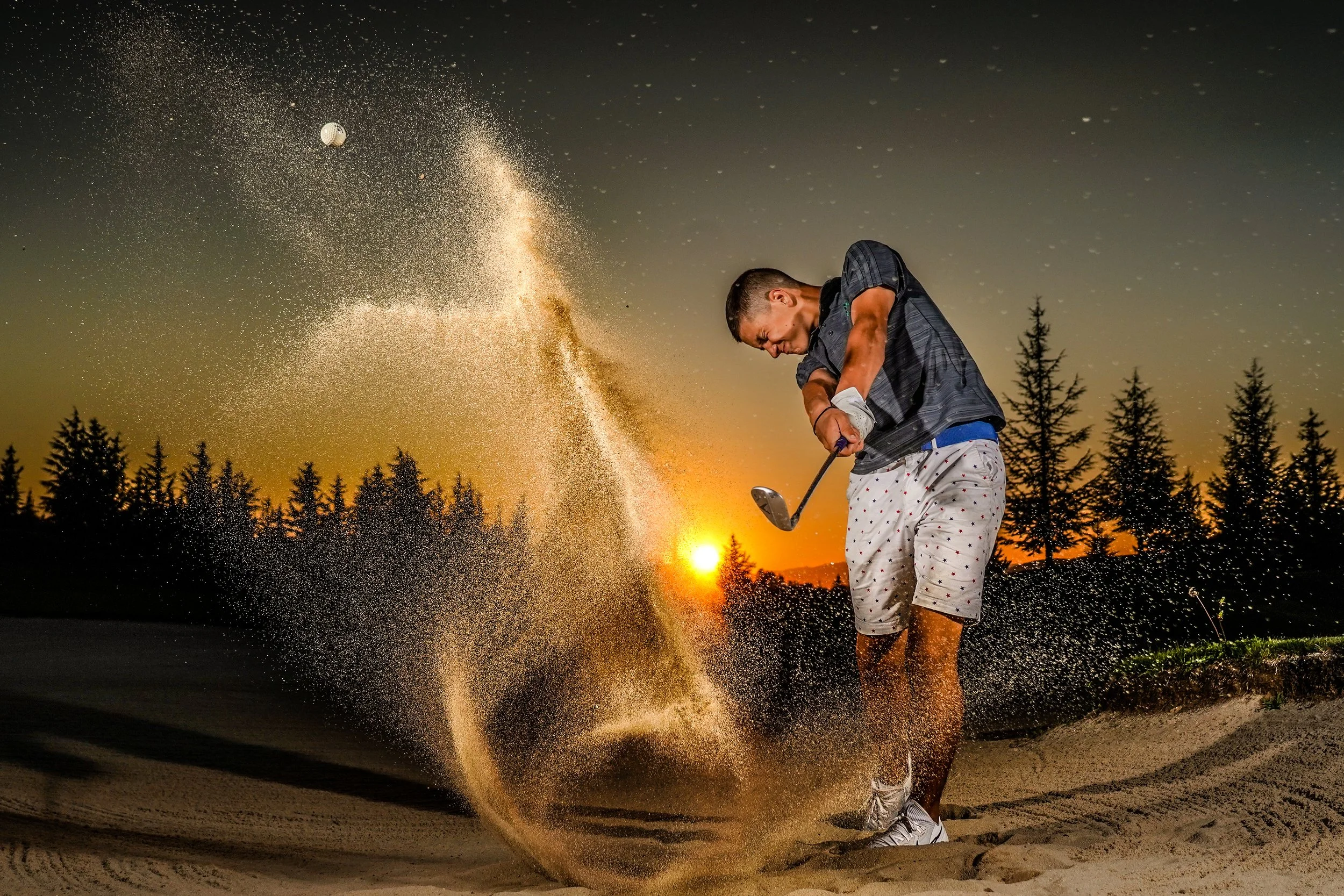 A man playing golf in a sand trap at sunset, swinging a club to hit a golf ball, with sand flying through the air and trees in the background.