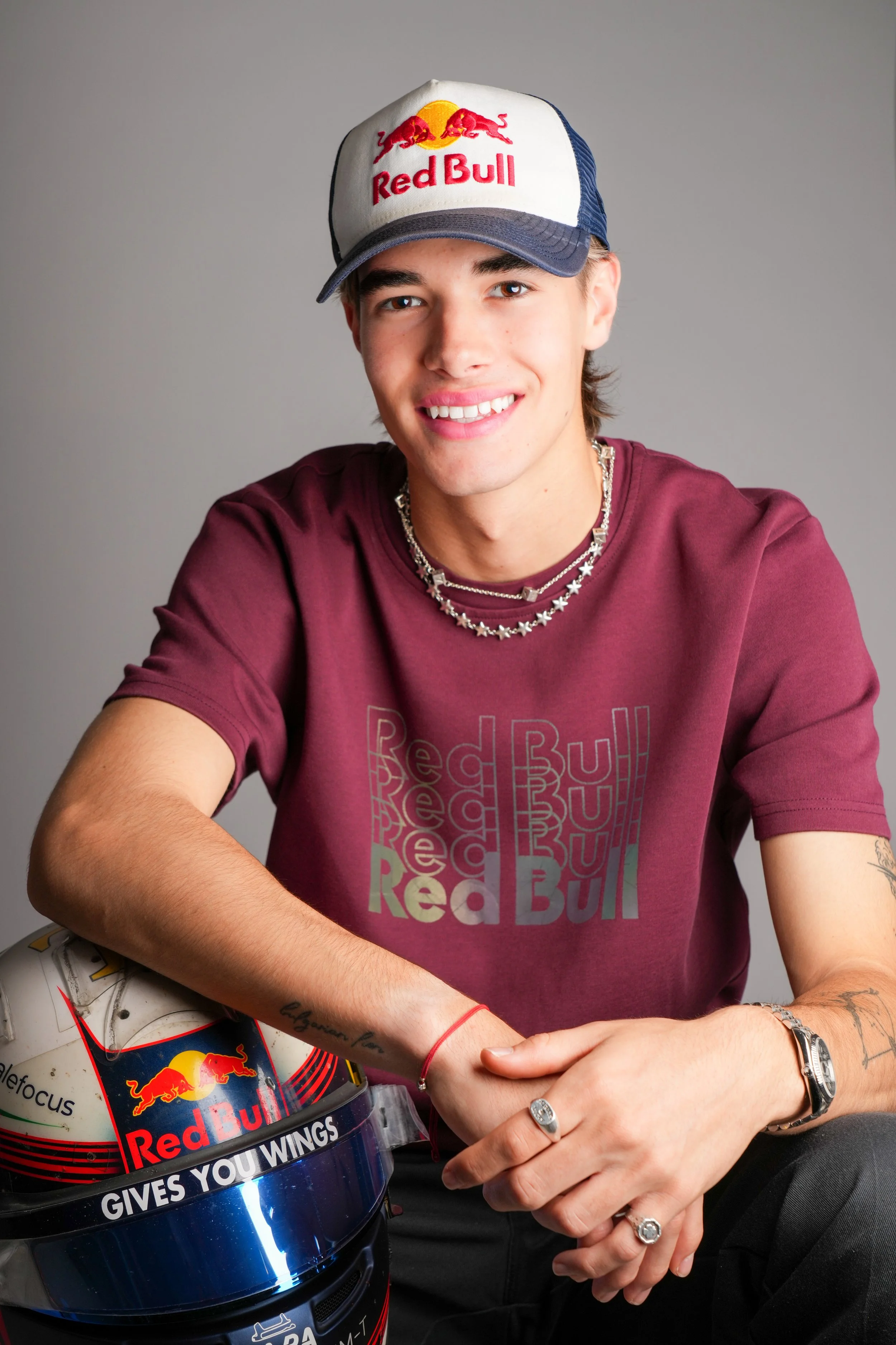 Young man wearing a Red Bull cap, maroon Red Bull t-shirt, and jewelry, sitting with a Red Bull helmet that has a Red Bull sticker.