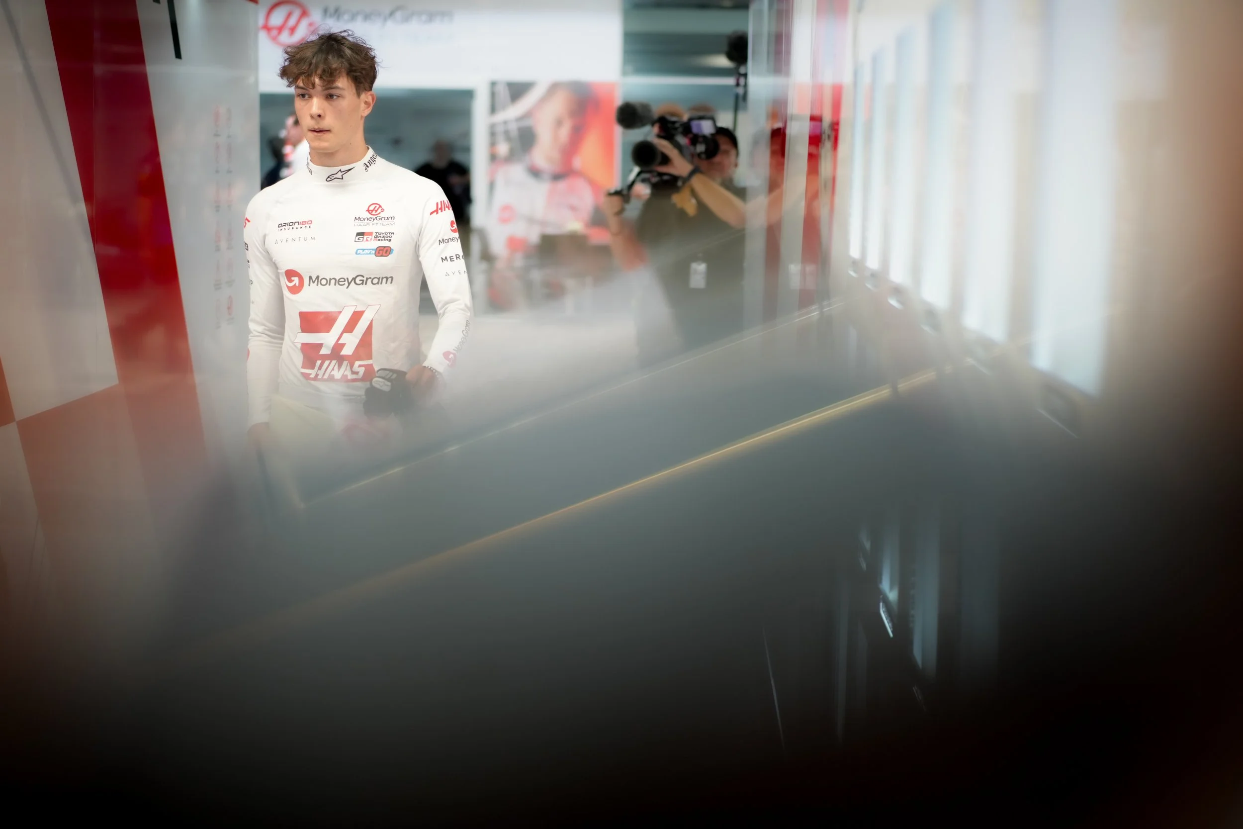 A male racing driver in a white Haas F1 team suit with logos, standing in the garage area, with a cameraman taking photos in the background.