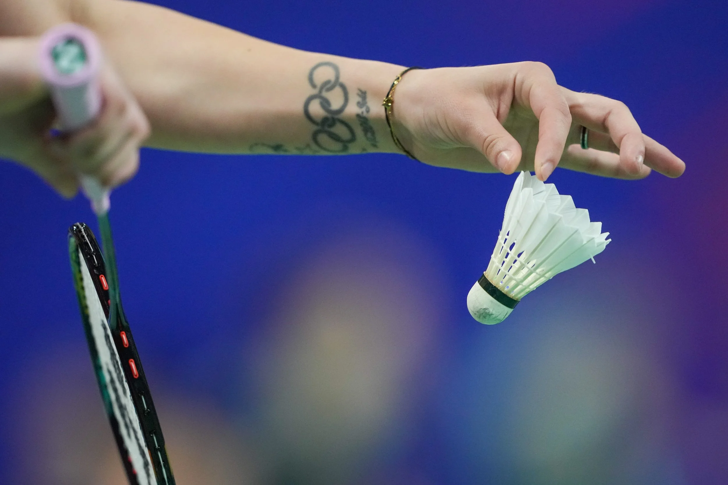 A person playing badminton, holding a badminton shuttlecock in one hand and a racket in the other, with a blue background.