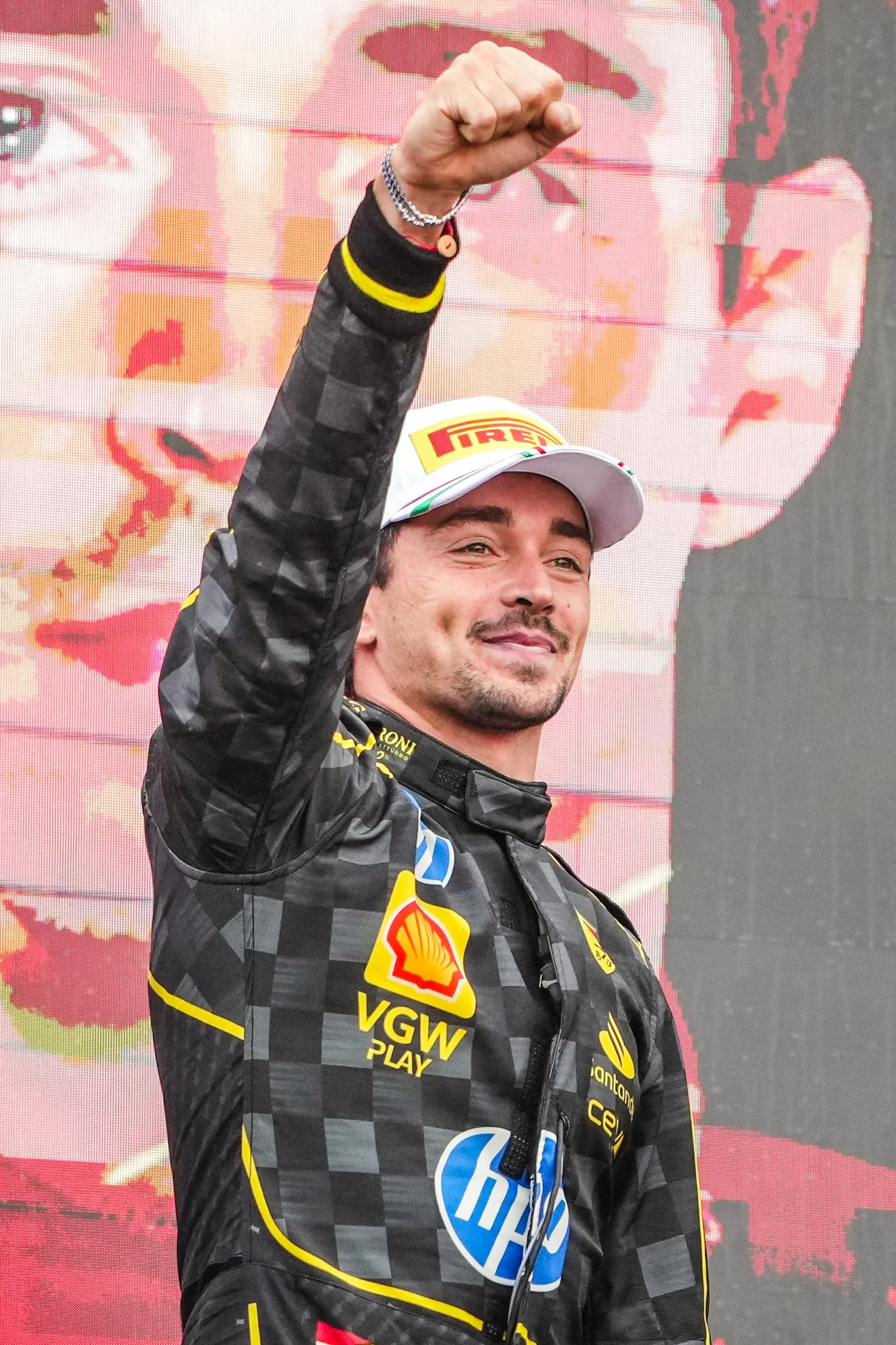 A man in racing attire with sponsor logos, wearing a white cap, raising his fist in victory, standing in front of a digital screen displaying a large image of himself.