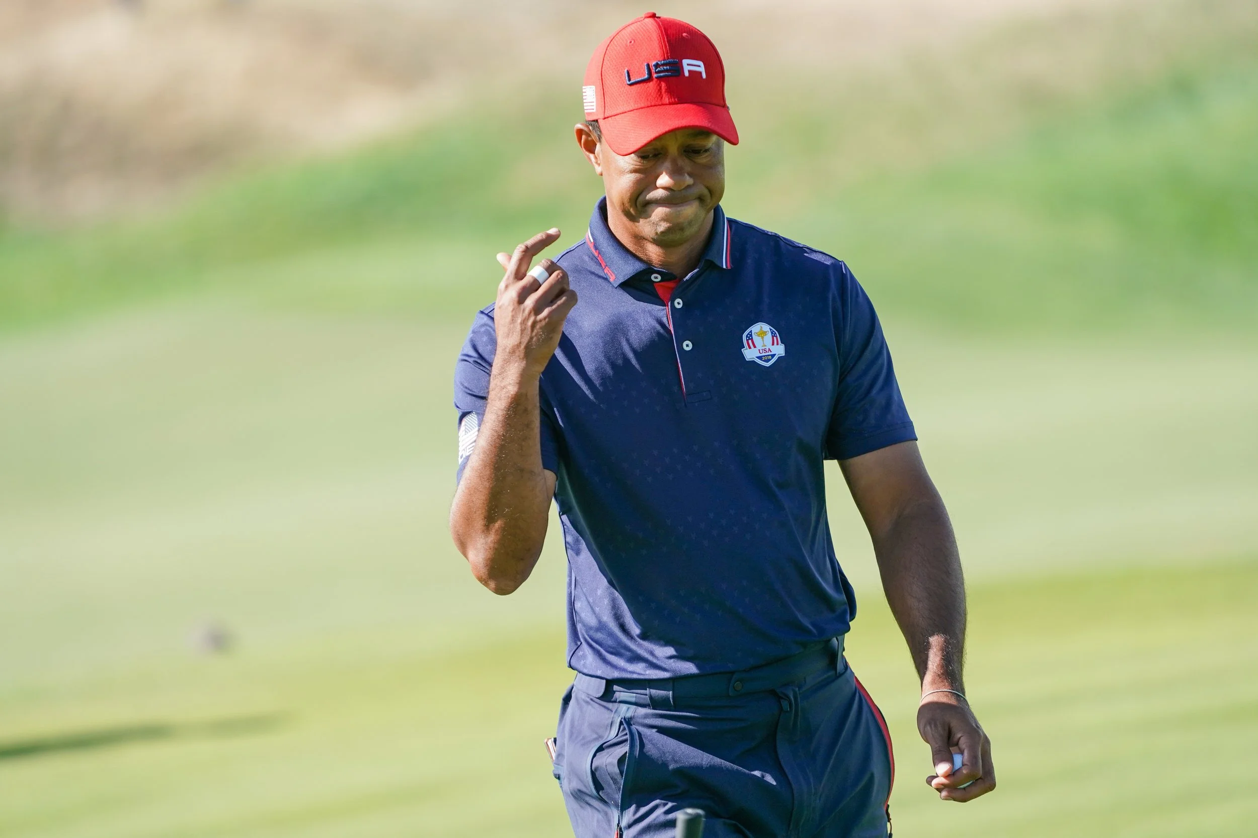 Man in navy blue polo shirt and red hat with USA logo standing on grassy field, holding golf club, with a focused expression.