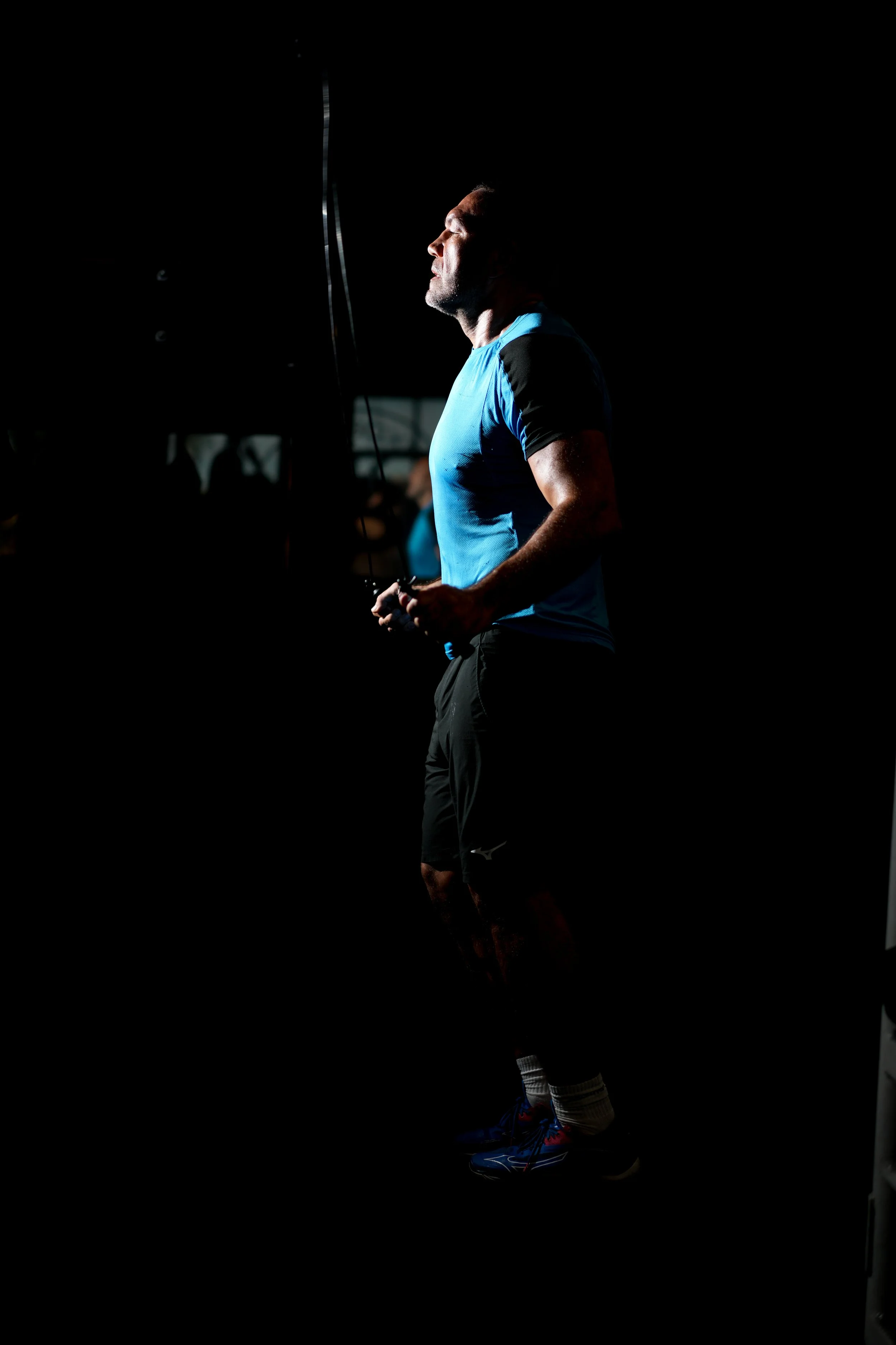 A man in a blue and black athletic shirt and black shorts holding a jump rope, standing in a dark gym with minimal lighting.