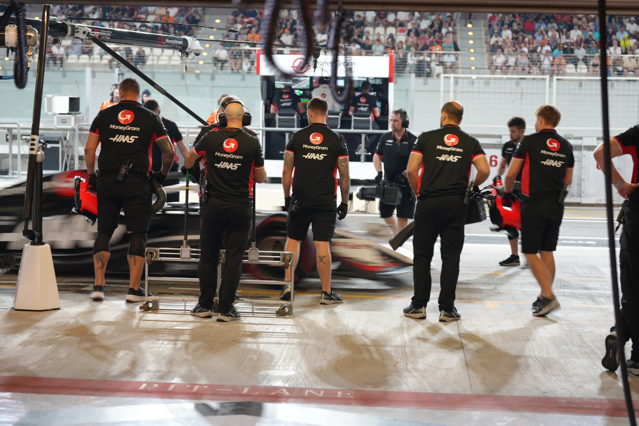F1 team members working in a pit stop garage with a blurred race car passing by, and a crowd of spectators in the stands in the background.