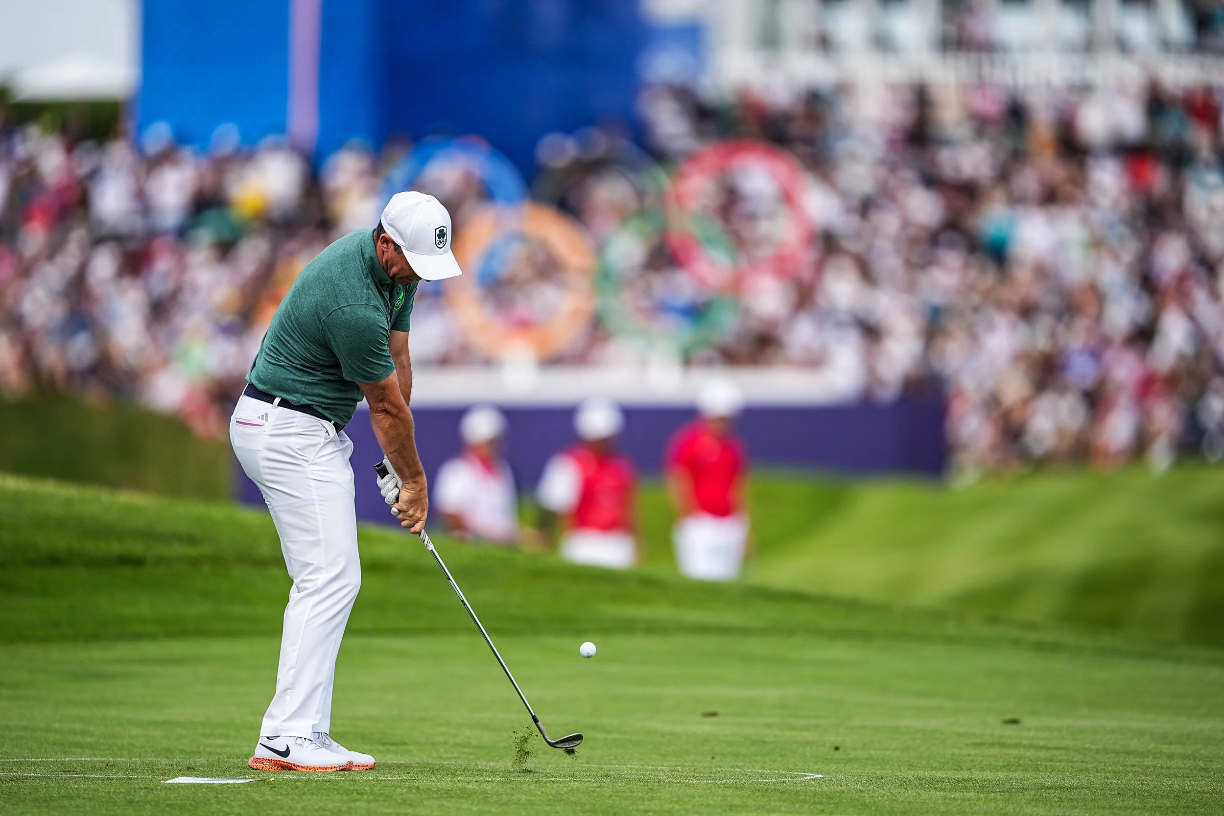A male golfer wearing a green shirt, white pants, and a white cap prepares to hit a golf ball on a putting green during a tournament, with a large crowd watching in the background.