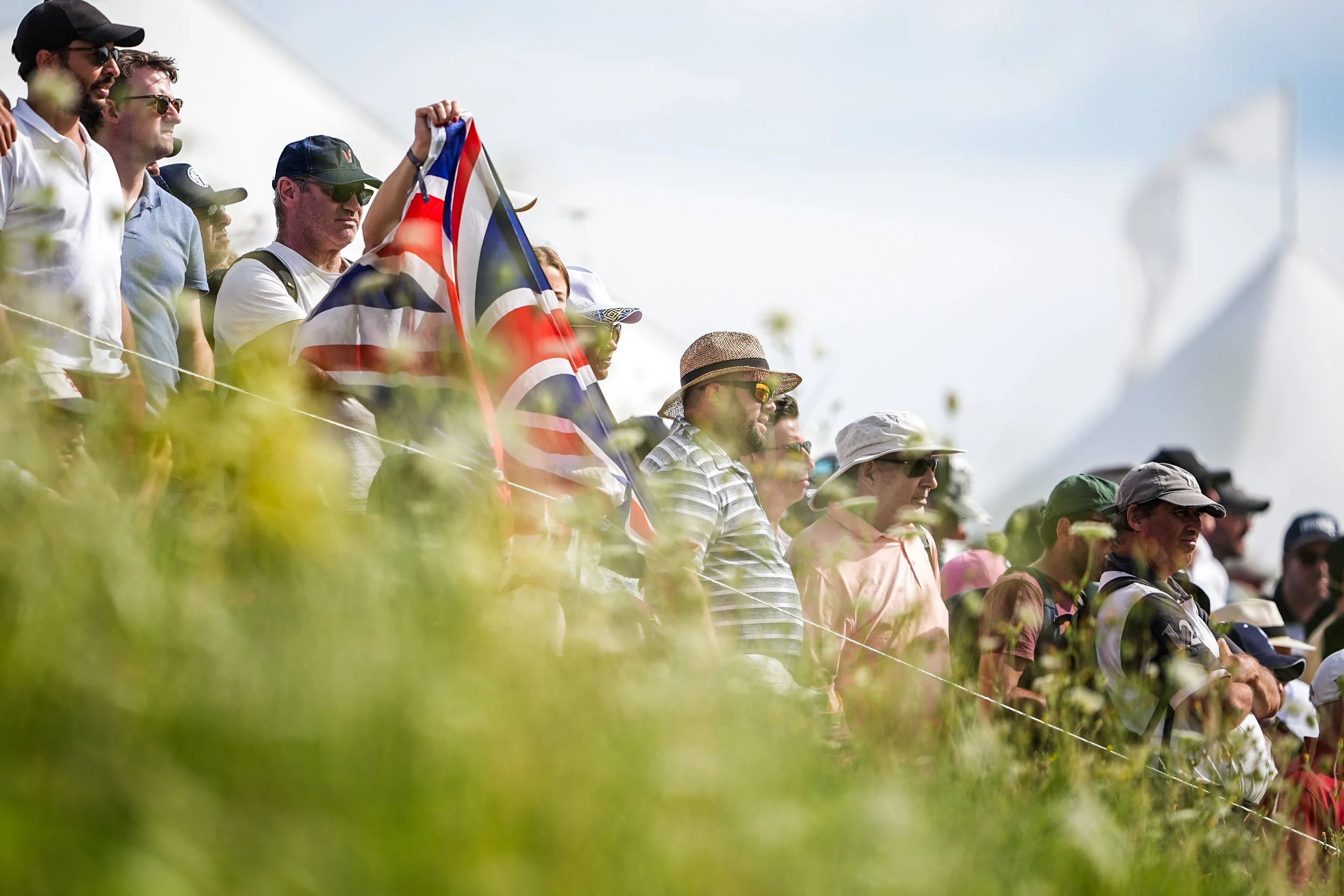 Crowd of people watching an outdoor event, some holding a Union Jack flag, with white tents in the background on a sunny day.