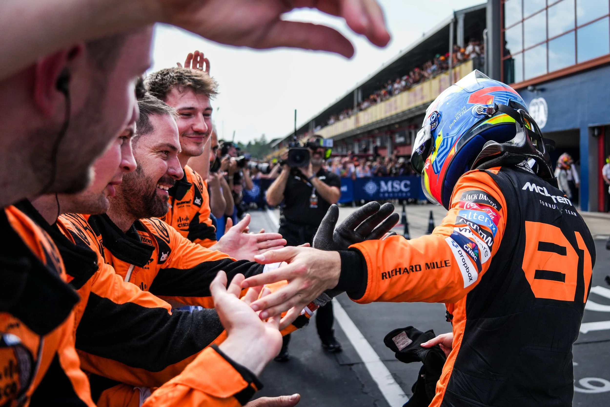 Formula 1 driver in racing gear and helmet interacting with team members in the pit lane, with spectators and team crew in the background.