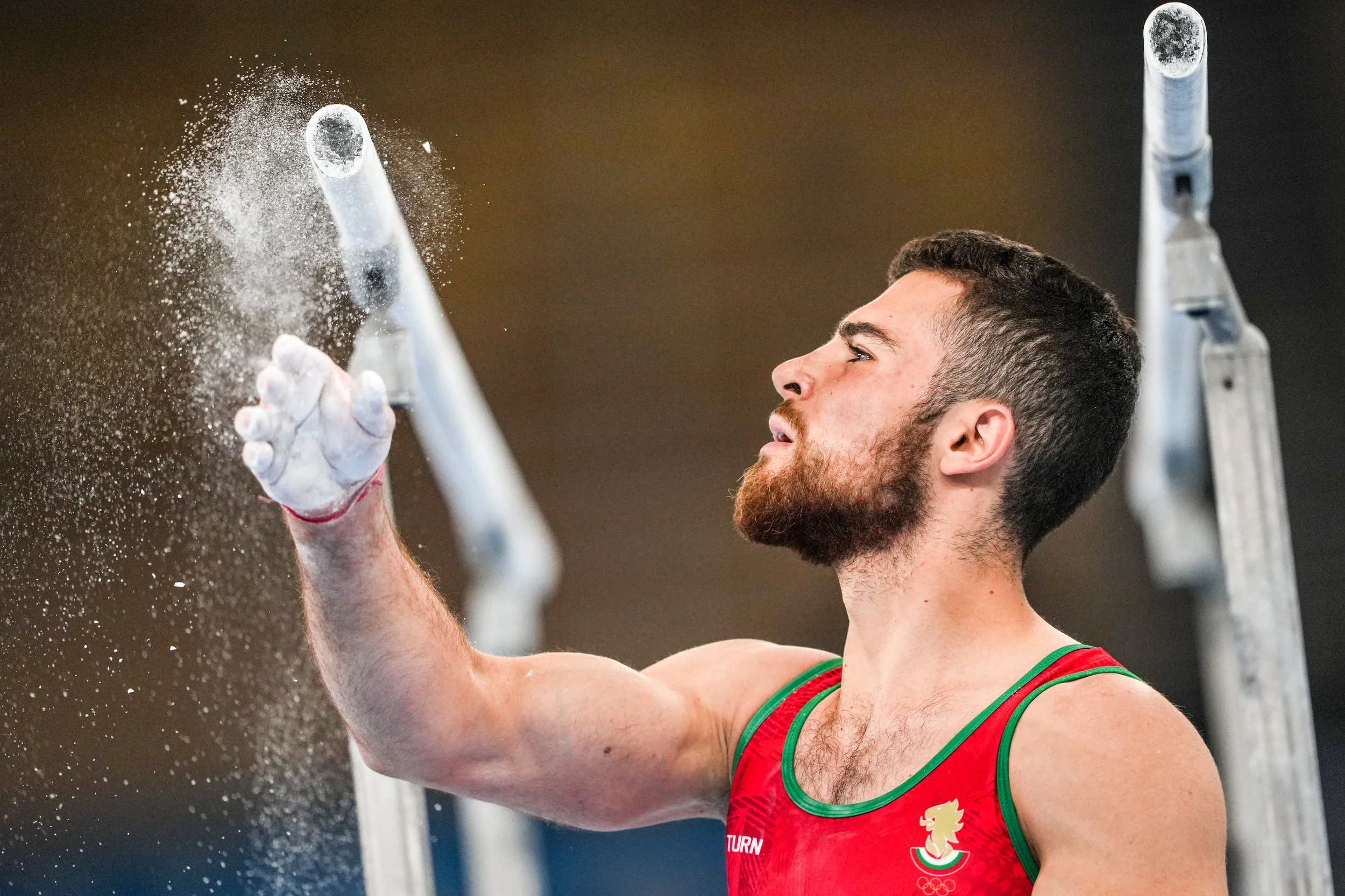 A male athlete in a red sports tank top with a graphic of a dragon and an Olympic logo, dusting chalk off his hand during a pole vault event. There are metal poles in the background.
