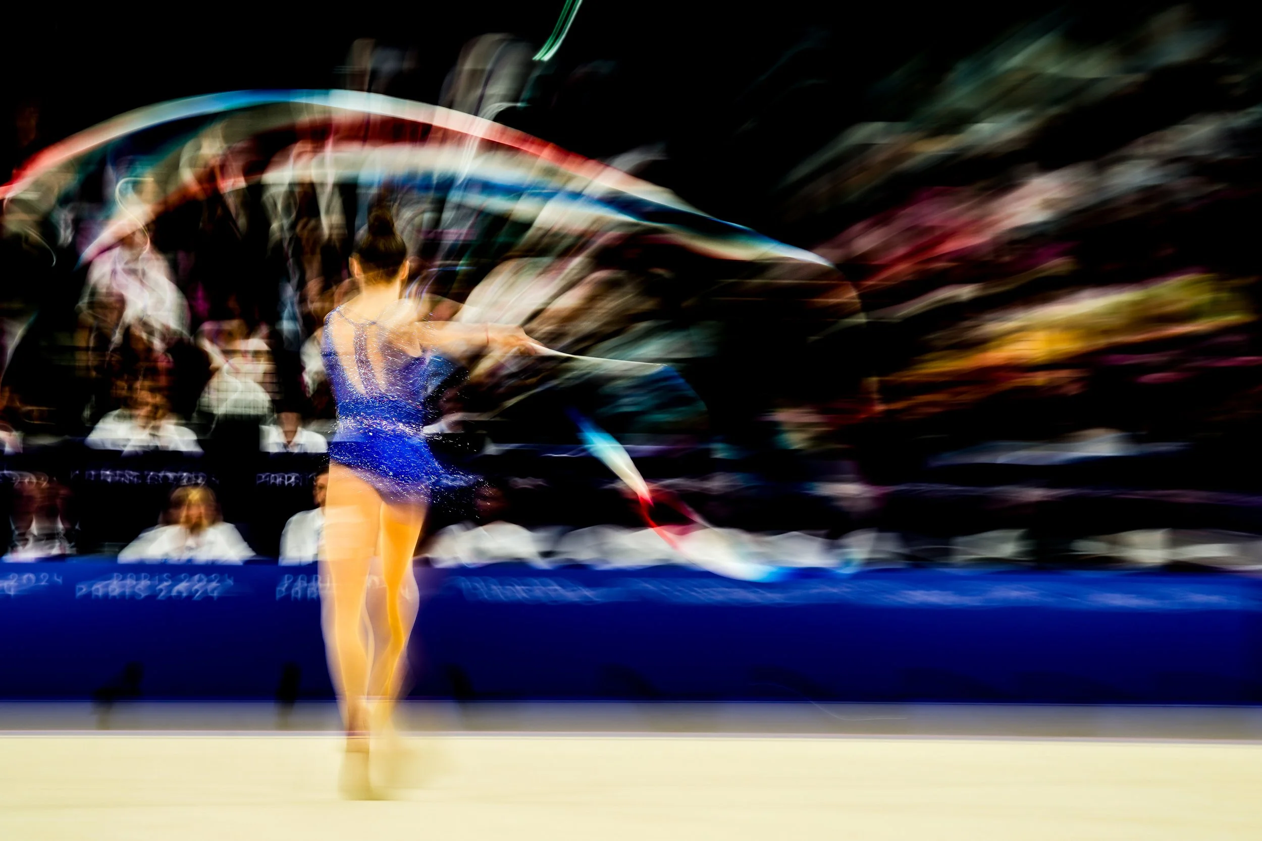 A gymnast in a blue costume performing with a ribbon in an indoor arena, with a blurred audience in the background.