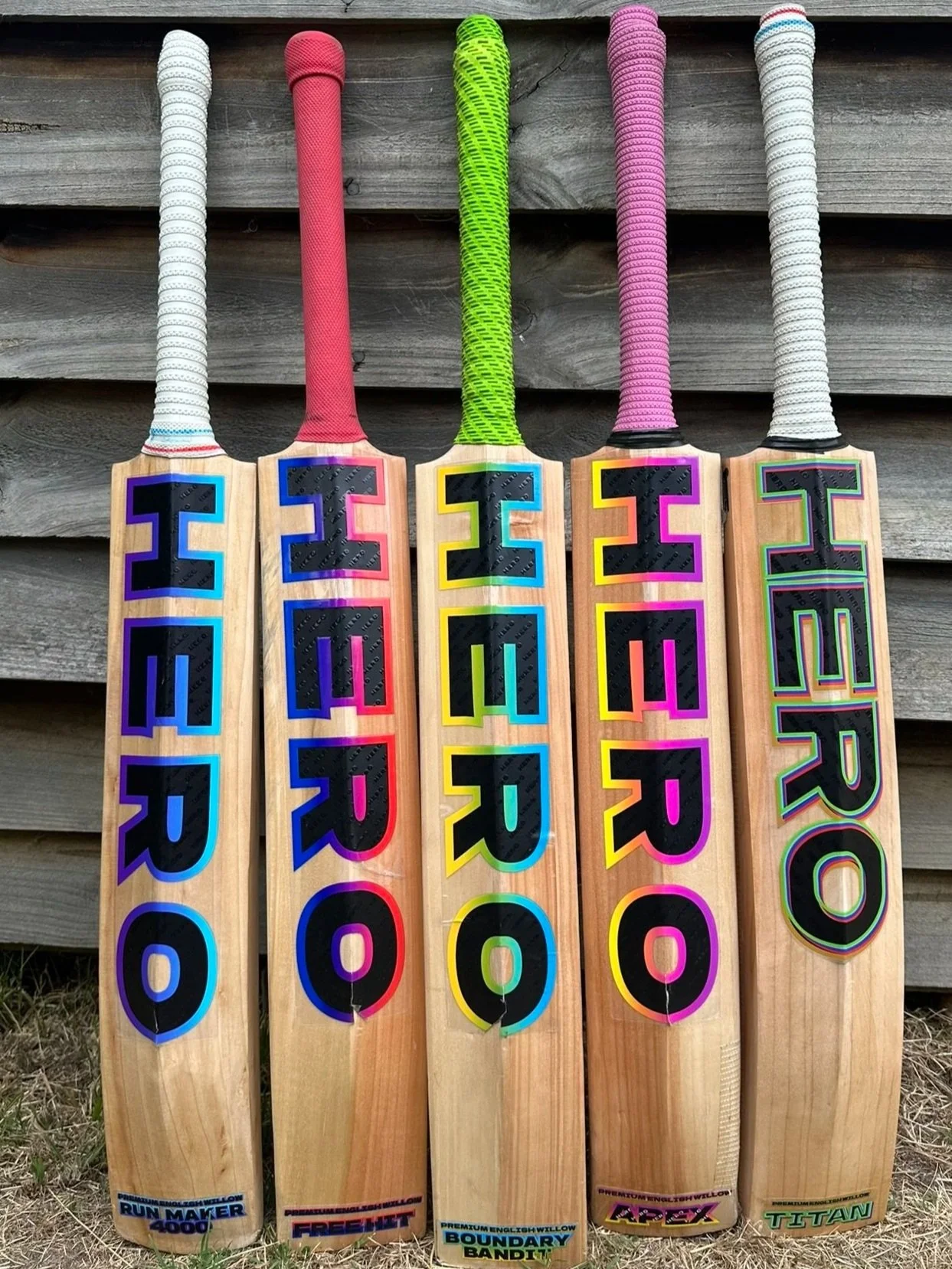 Five cricket bats with colorful handles and large black letters spelling 'HERO' on each bat, leaning against a wooden fence.
