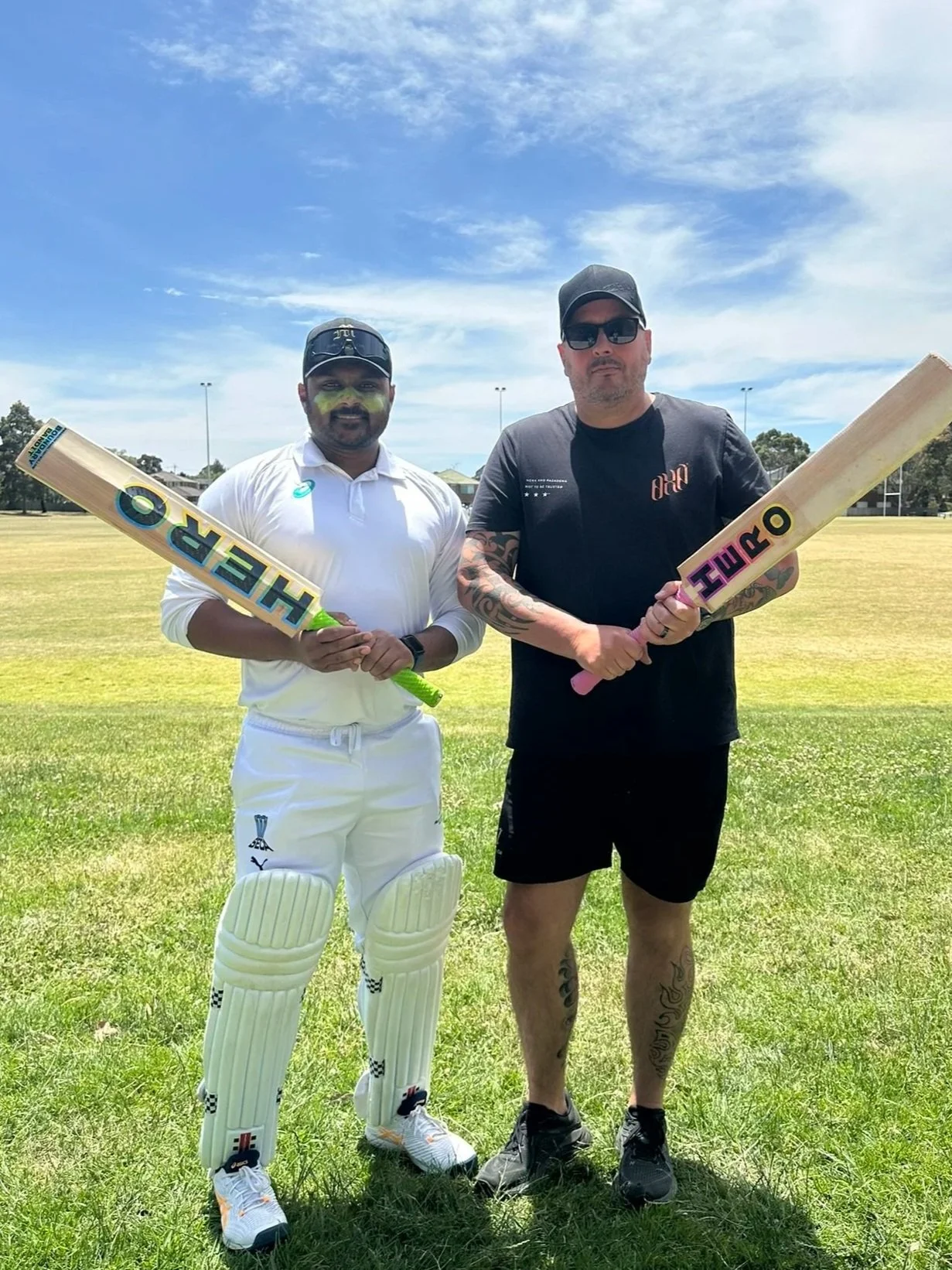 Two men standing on a grassy cricket field holding cricket bats. One man is in cricket gear and the other in casual black clothing and sunglasses. Clear blue sky overhead.