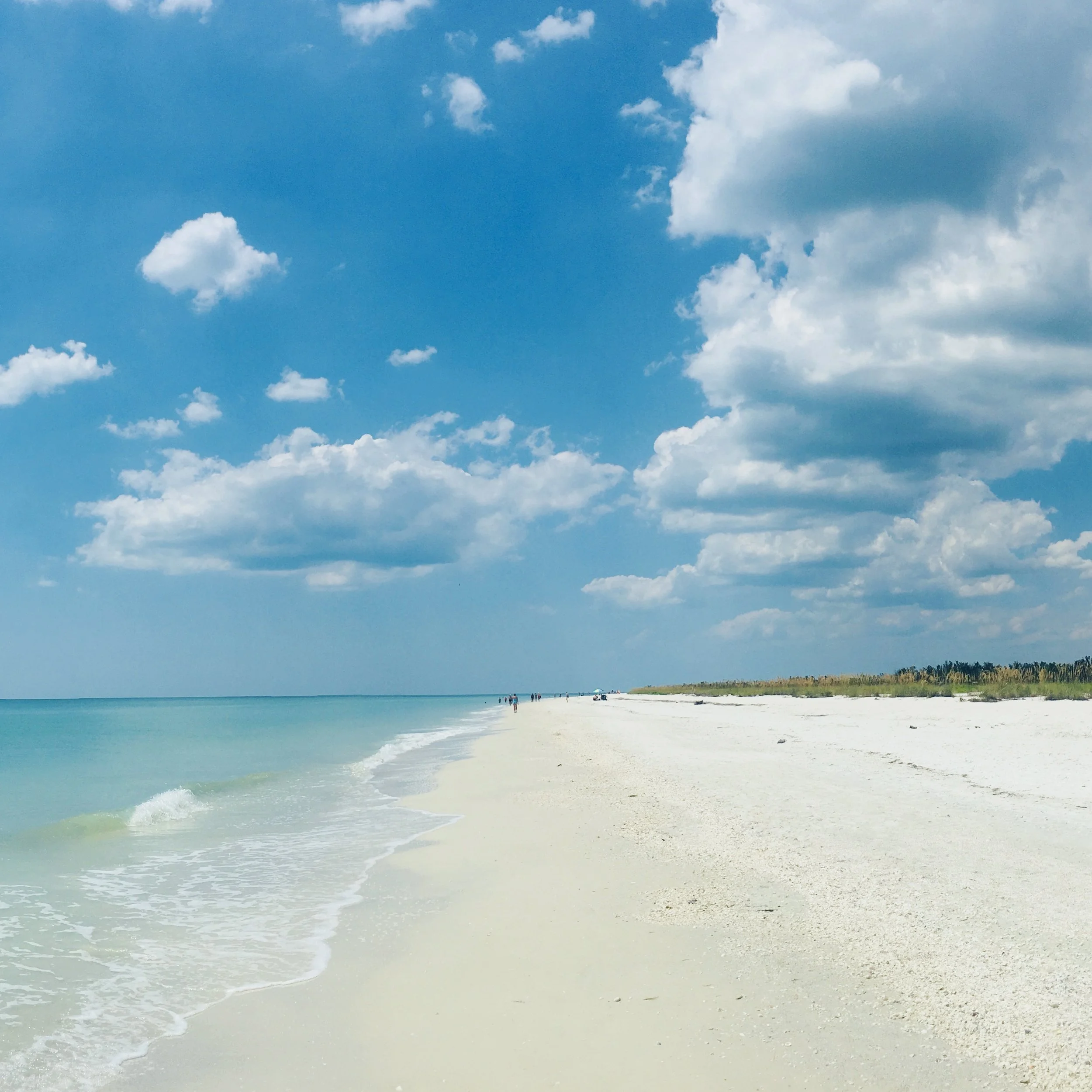 Ein ruhiger Strand mit weißem Sand, ruhigem Meer und einigen Menschen, die am Wasser entlang gehen, bei klarem Himmel mit wenigen Wolken.