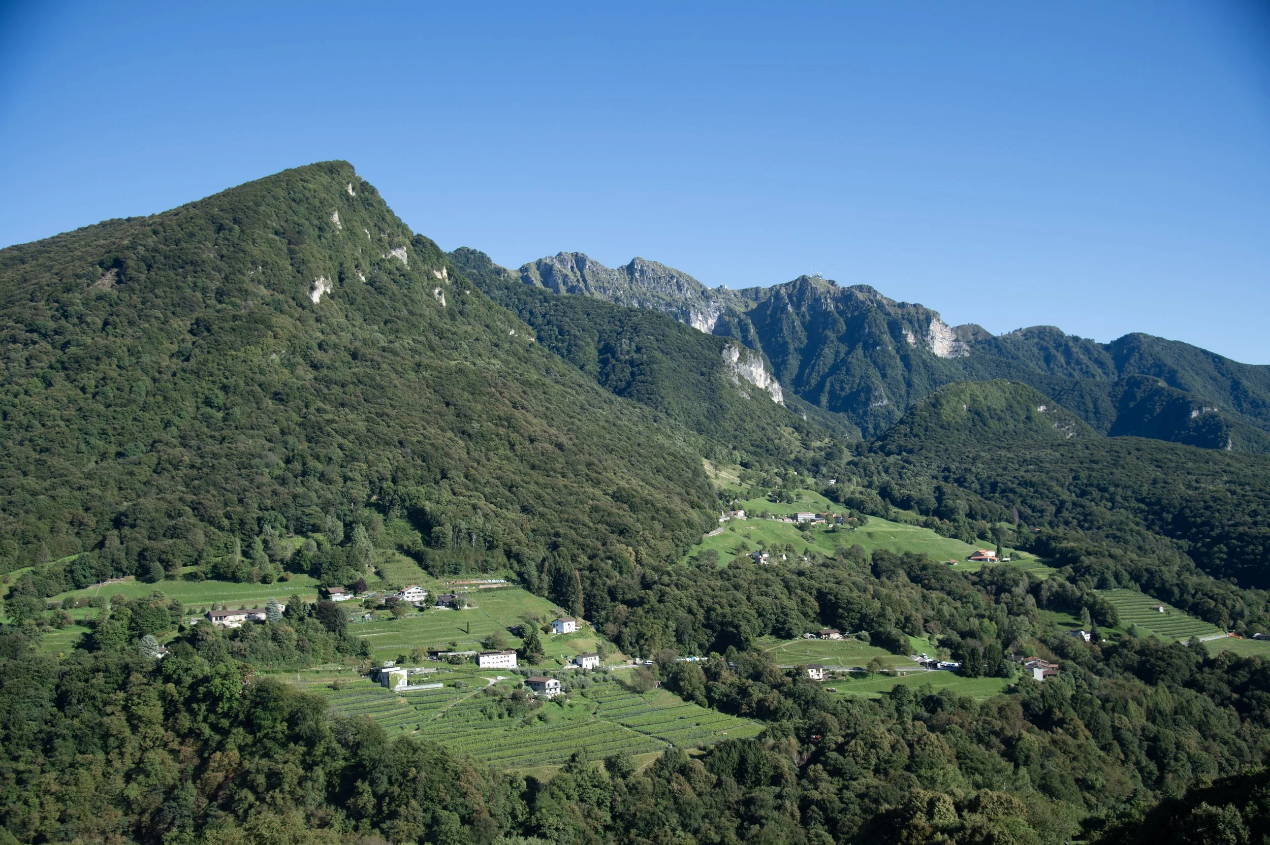 Paesaggio montano con colline coltivate e case sparse, con montagne verdi sullo sfondo e un cielo blu chiaro.