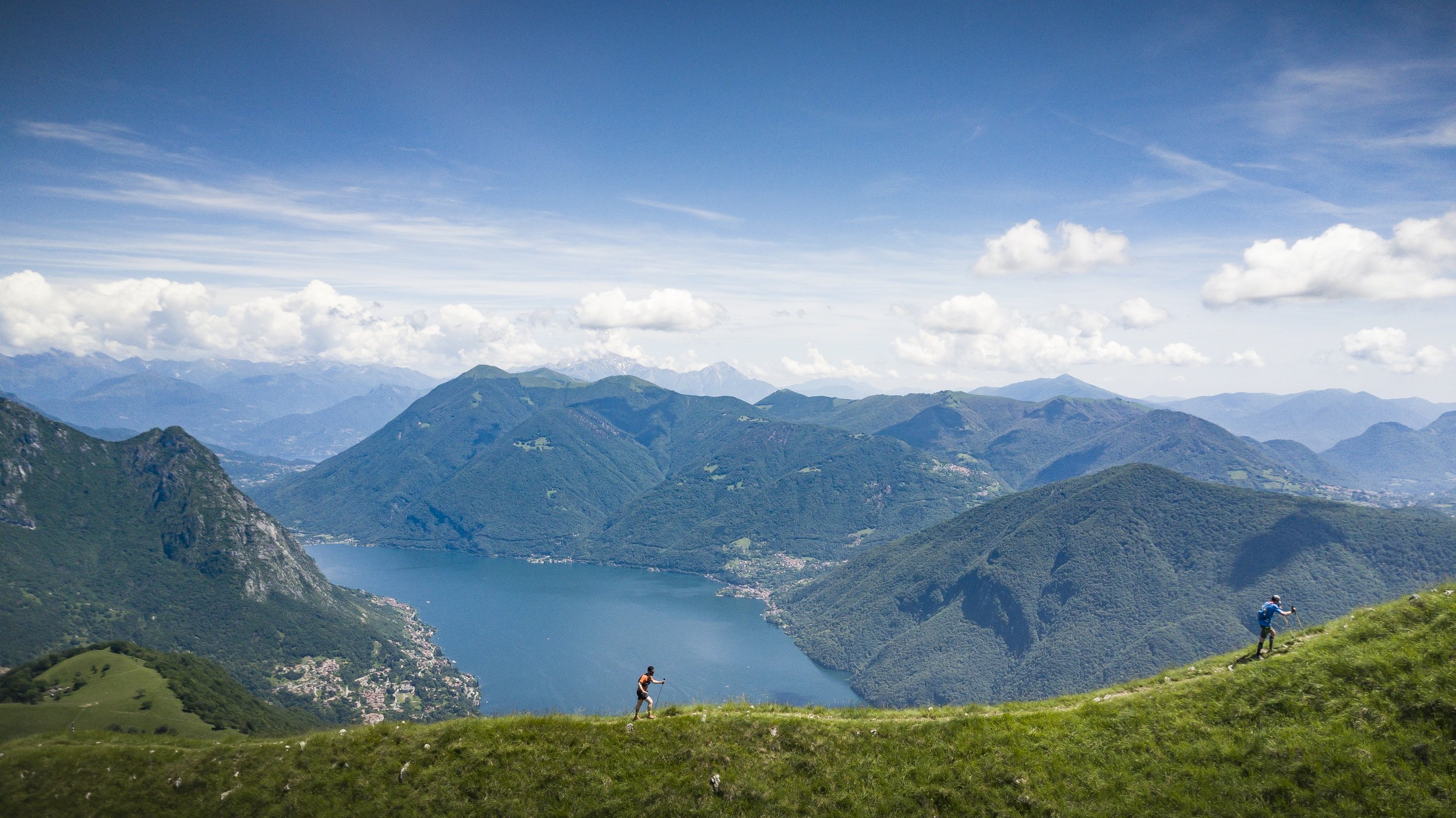 Due persone che fanno escursioni in un paesaggio montano con un grande lago, colline verdi e montagne sullo sfondo sotto un cielo azzurro con nuvole.