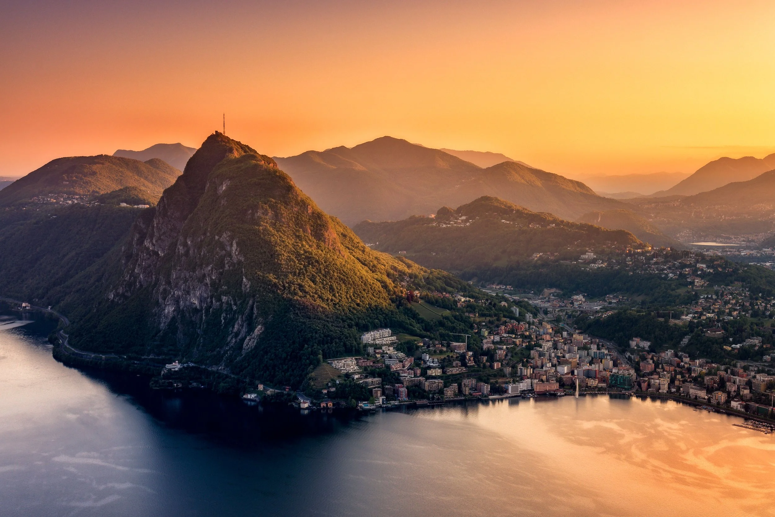 Vista panoramica di un lago circondato da città e montagne al tramonto, con una delle montagne più prominenti al centro