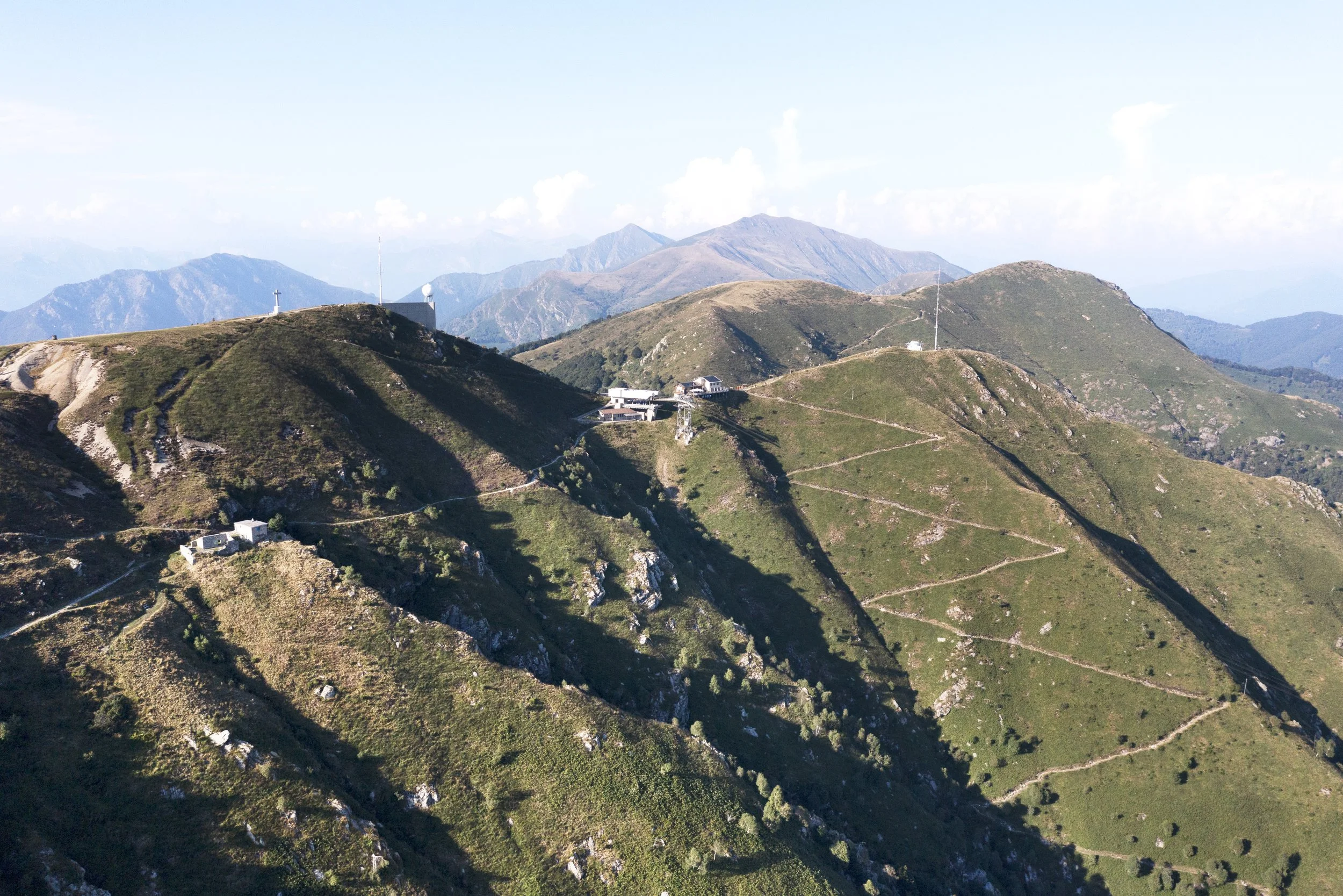 Paesaggio montano con sentieri che attraversano le cime verdi e alcune strutture su una delle montagne, con sfondo di altre montagne e cielo sereno.