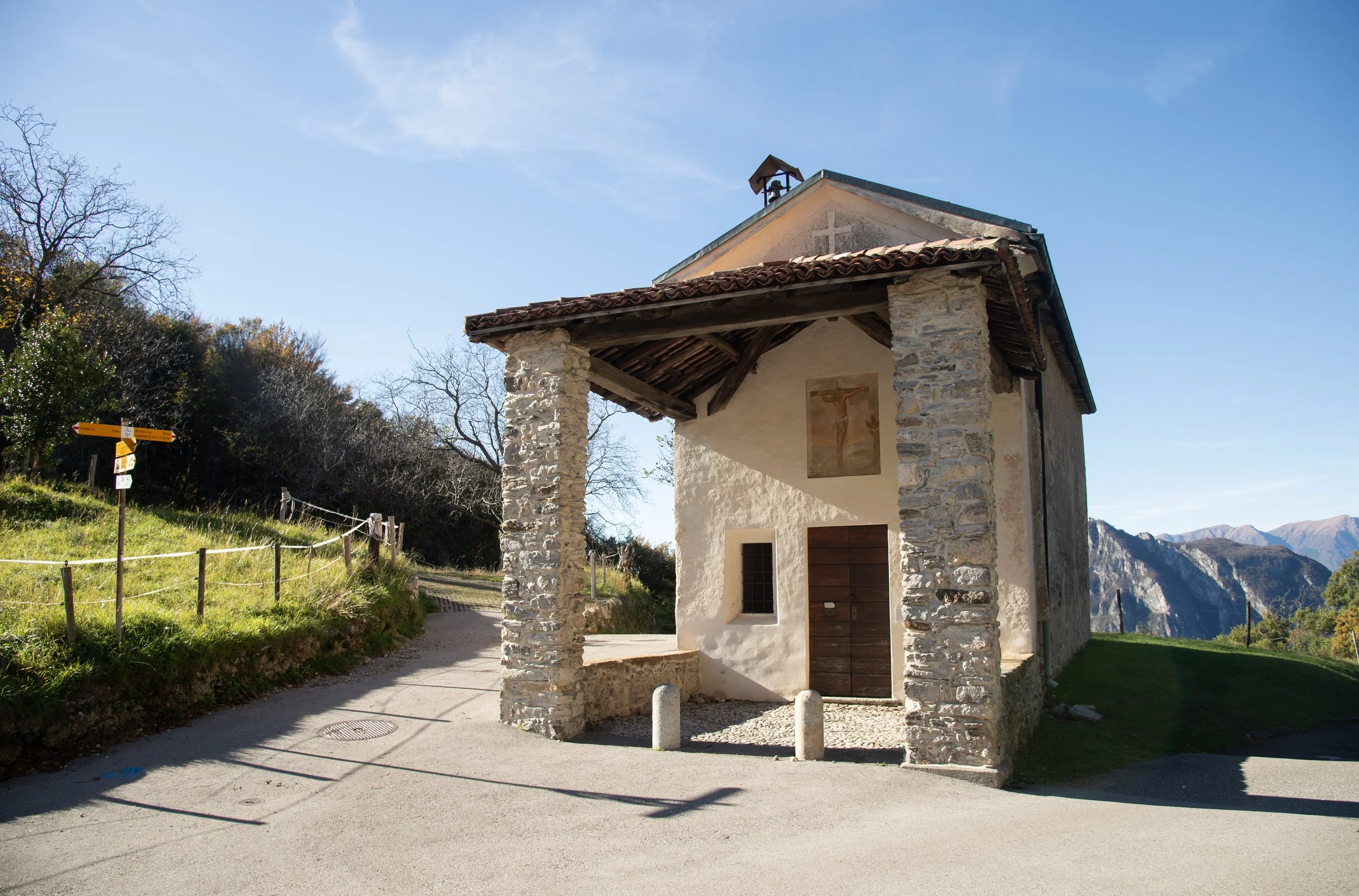 Chiesa di montagna in pietra con dipinto di Gesù crocifisso sulla parete esterna, su strada sterrata, con montagne sullo sfondo e cielo blu.