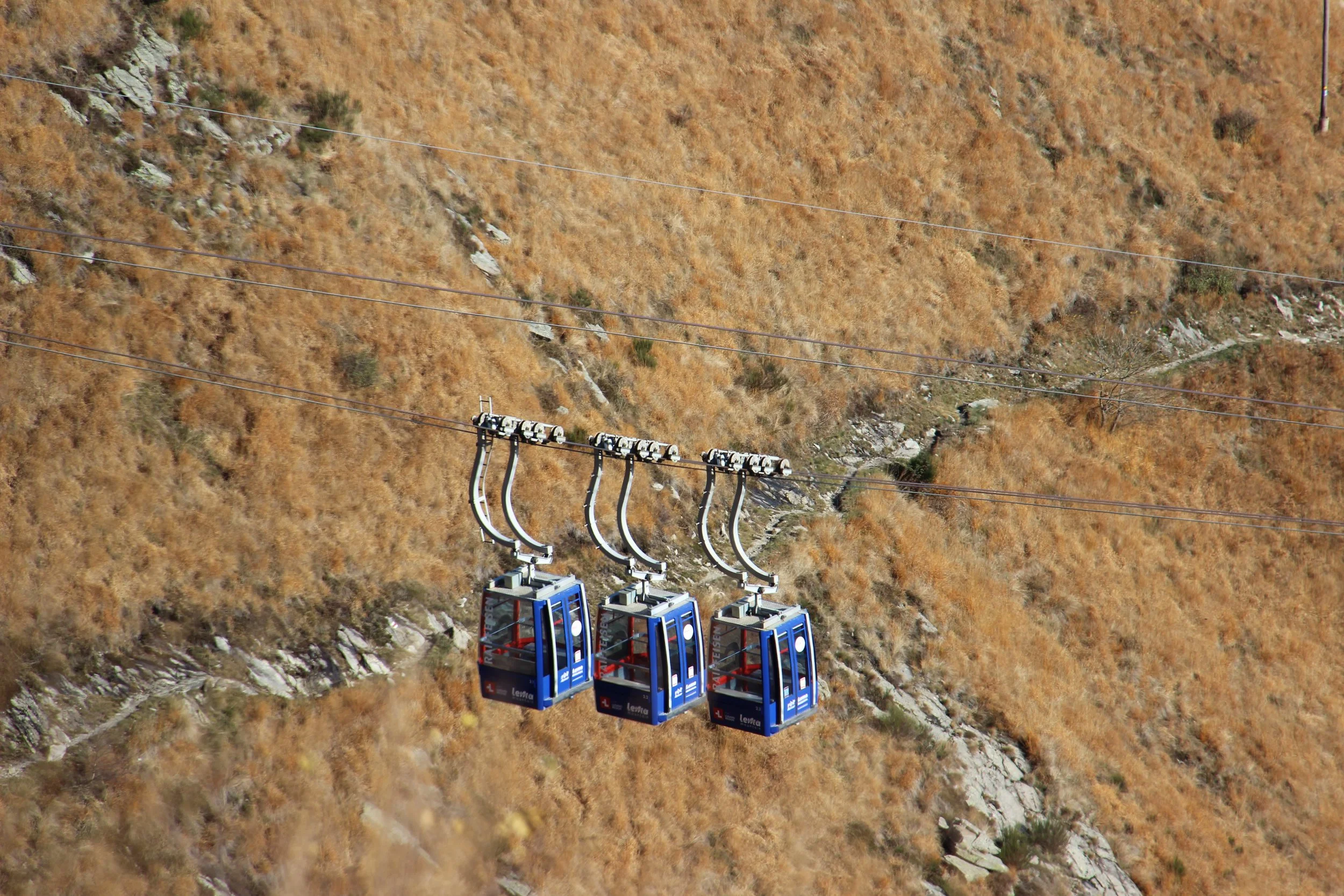 Tre cabine di un sistema di funivia blu sospese sopra un paesaggio di terra arida e rocciosa.