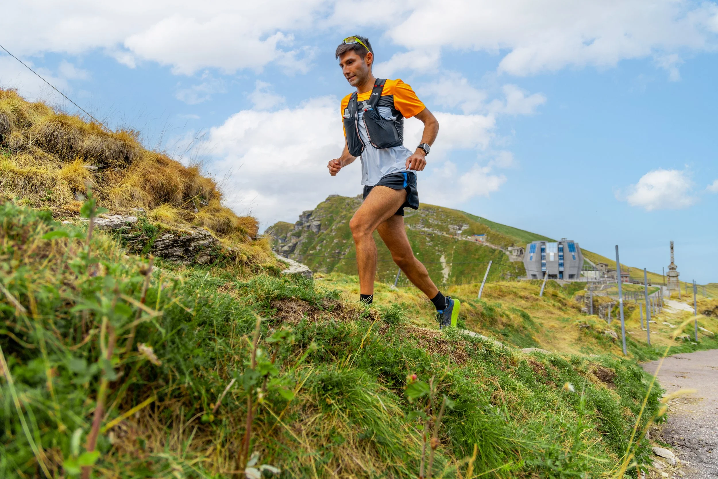 Uomo che fa trail running sulla costa, con colline verdi e un edificio moderno sullo sfondo, cielo nuvoloso.