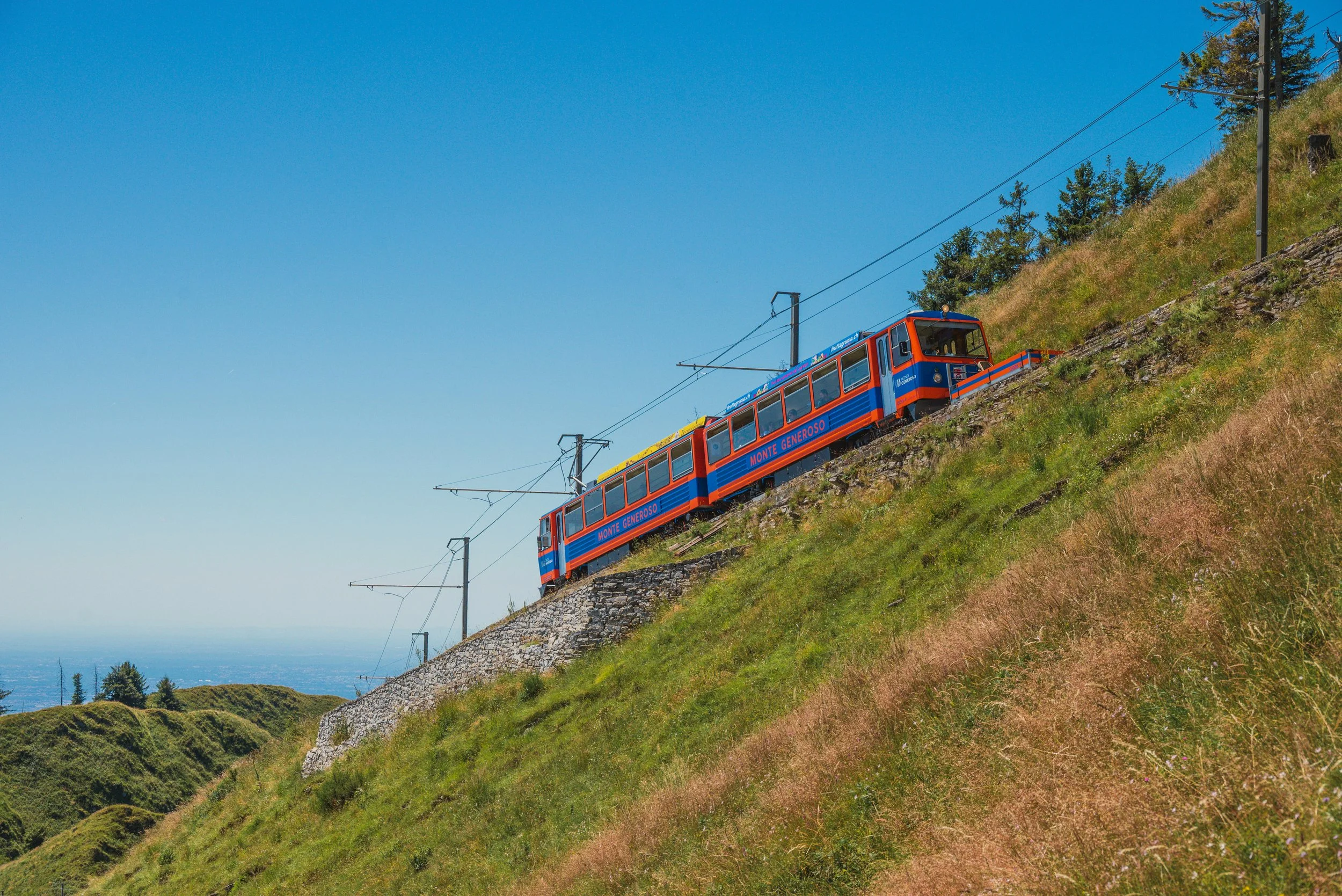 Il treno Monte Generoso con carrozze blu e arancioni percorre una linea ferroviaria inclinata sopra una collina verde.