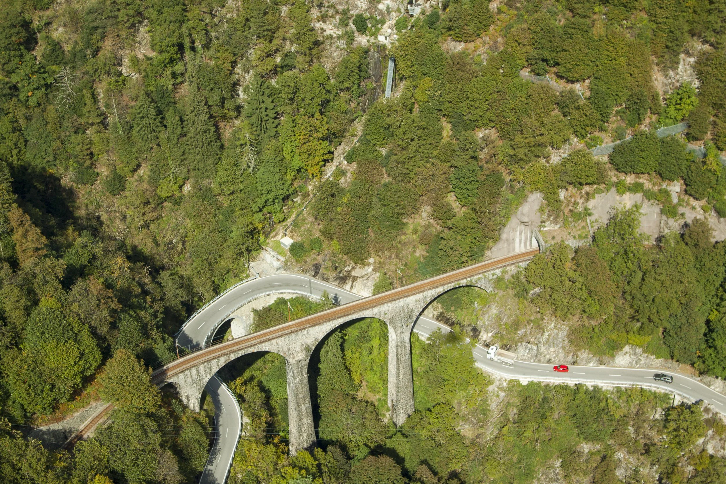 Vista aerea di un ponte in pietra che attraversa una valle montagnosa ricoperta di alberi verdi, con alcune curve sulla strada