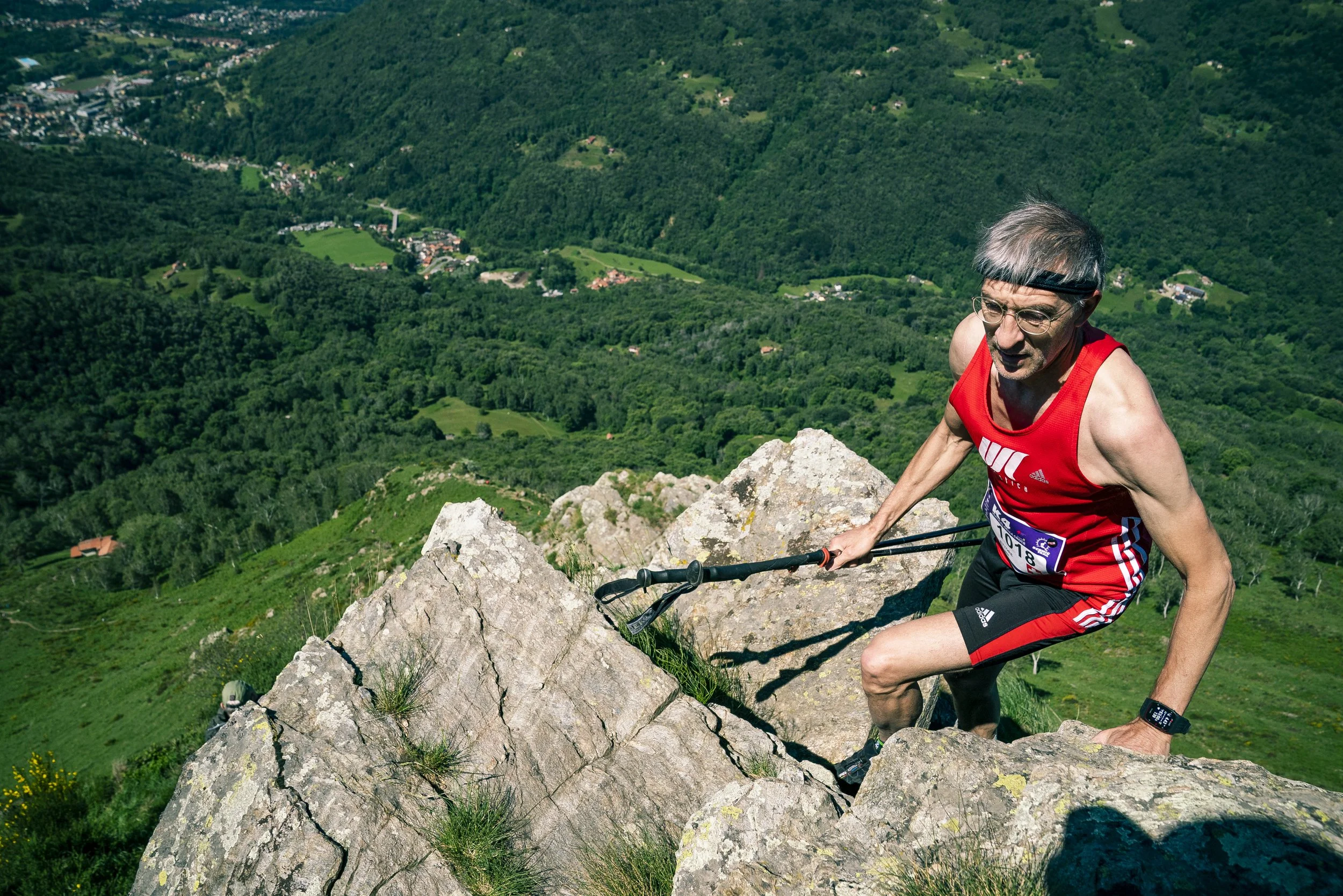 Uomo anziano che arrampica una parete di rocce in montagna durante una corsa o una gara, con un paesaggio verde e montuoso sullo sfondo.