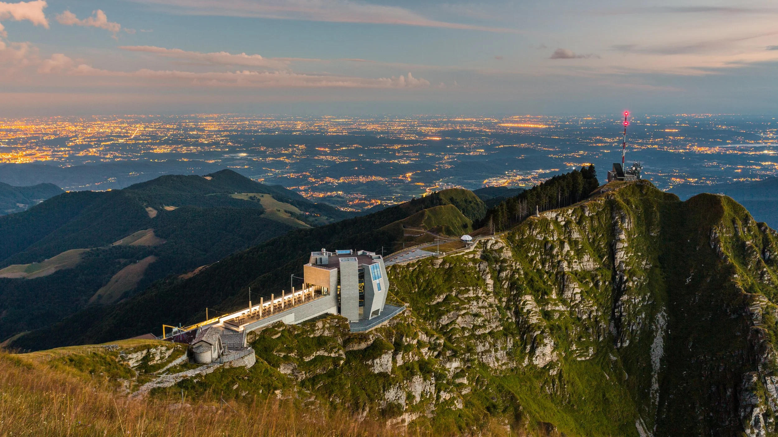 Vista serale delle montagne con un edificio moderno e un'antenna a lancia sulla cima, con luci della città in lontananza.