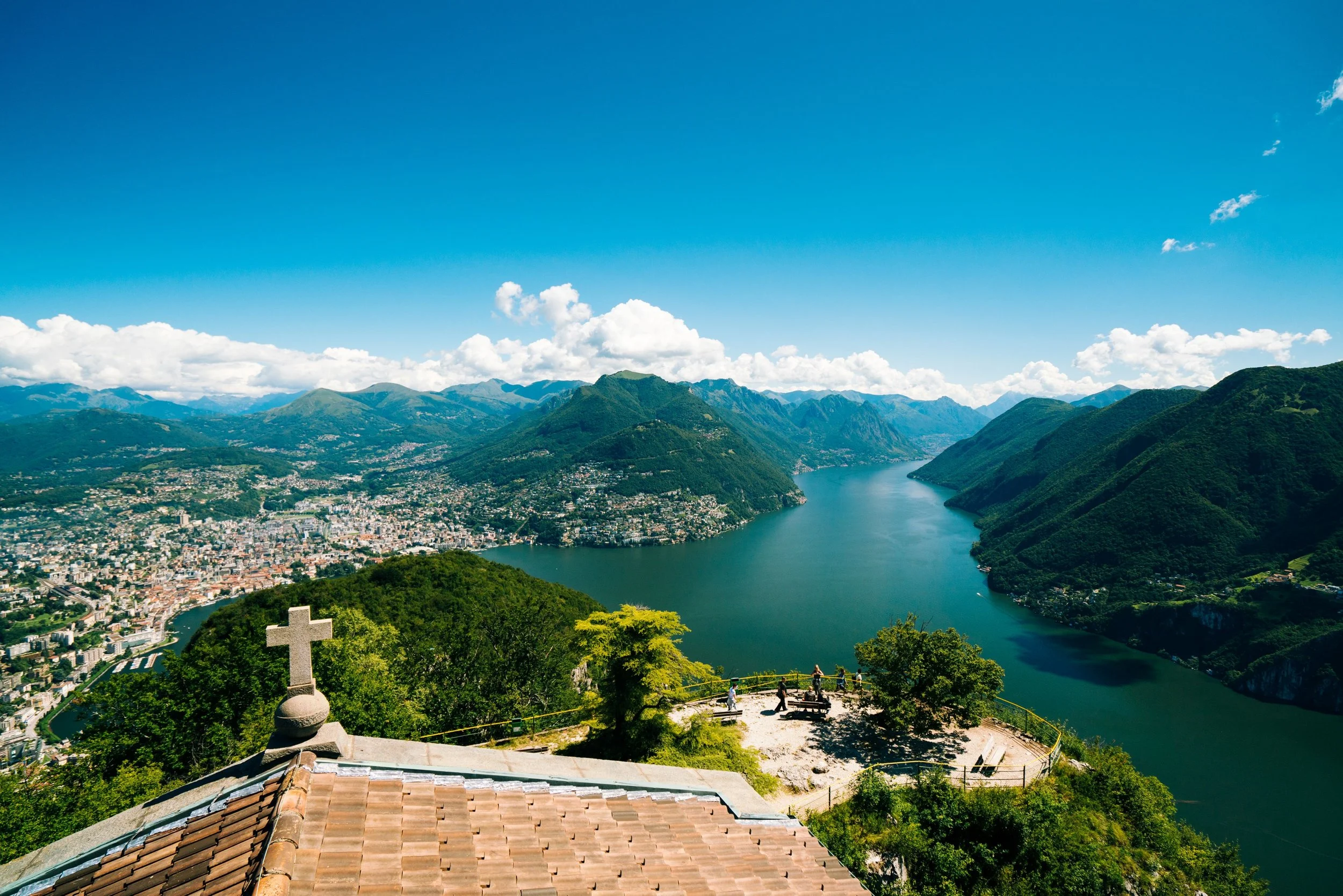 Paesaggio naturale con lago, montagne, cielo azzurro e nuvole, vista dalla sommità di un punto panoramico con croce e panchine.