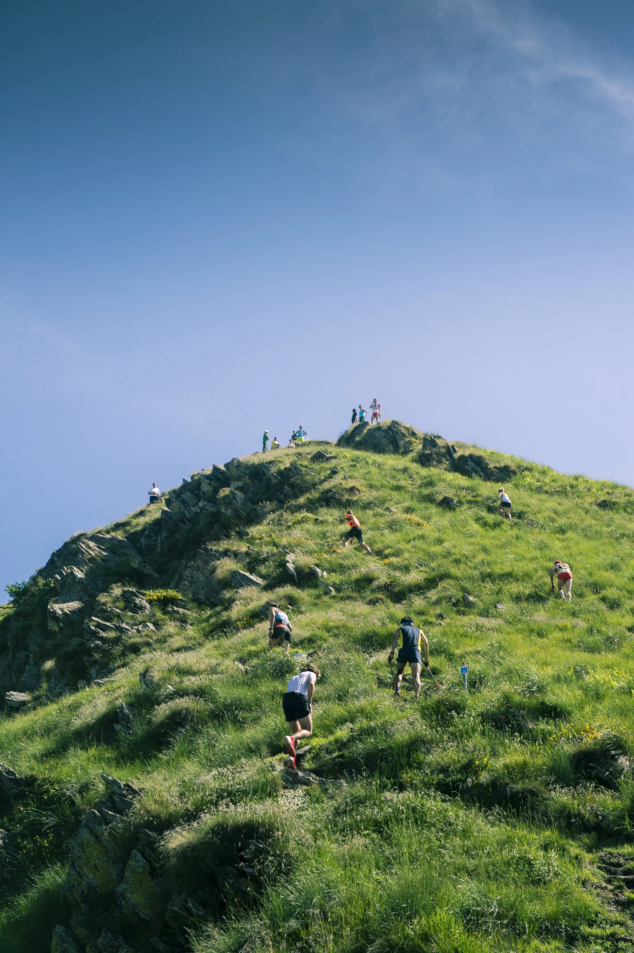 Un gruppo di persone che scalano una collina verde con rocce sotto un cielo azzurro.