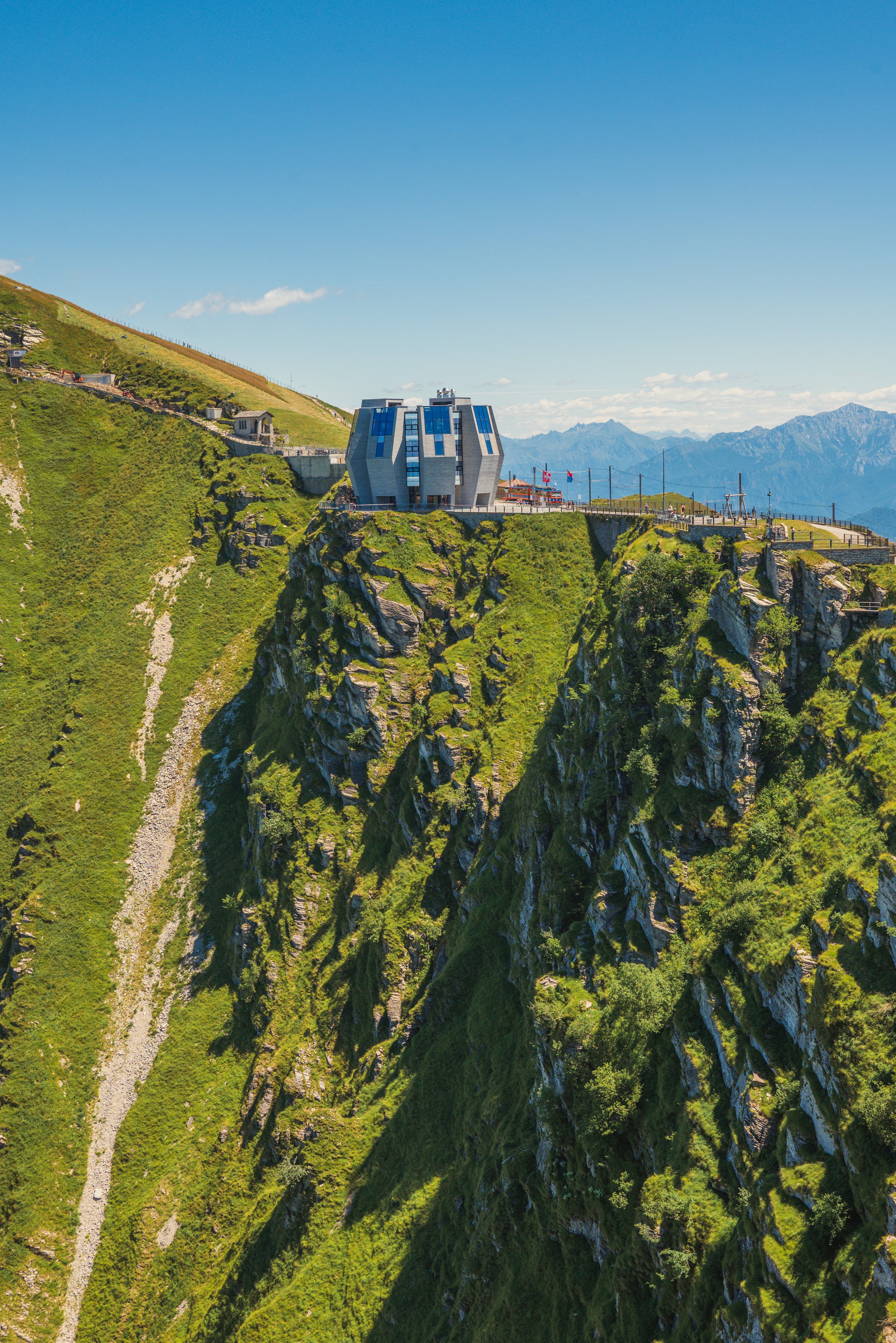 Edificio moderno sulla cima di una montagna verde e rocciosa sotto un cielo blu. Paesaggio montano in lontananza.