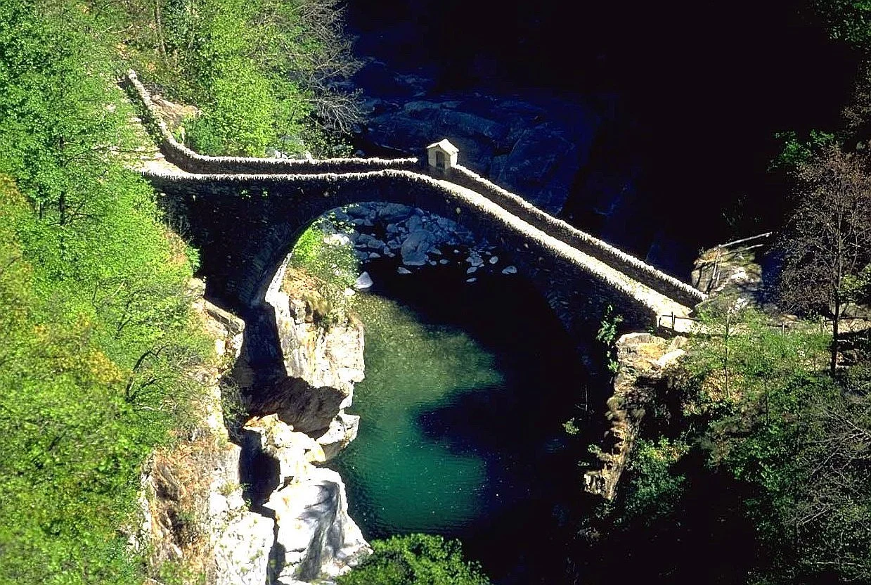 Ponte in pietra che attraversa un fiume in una zona verde con alberi e rocce.