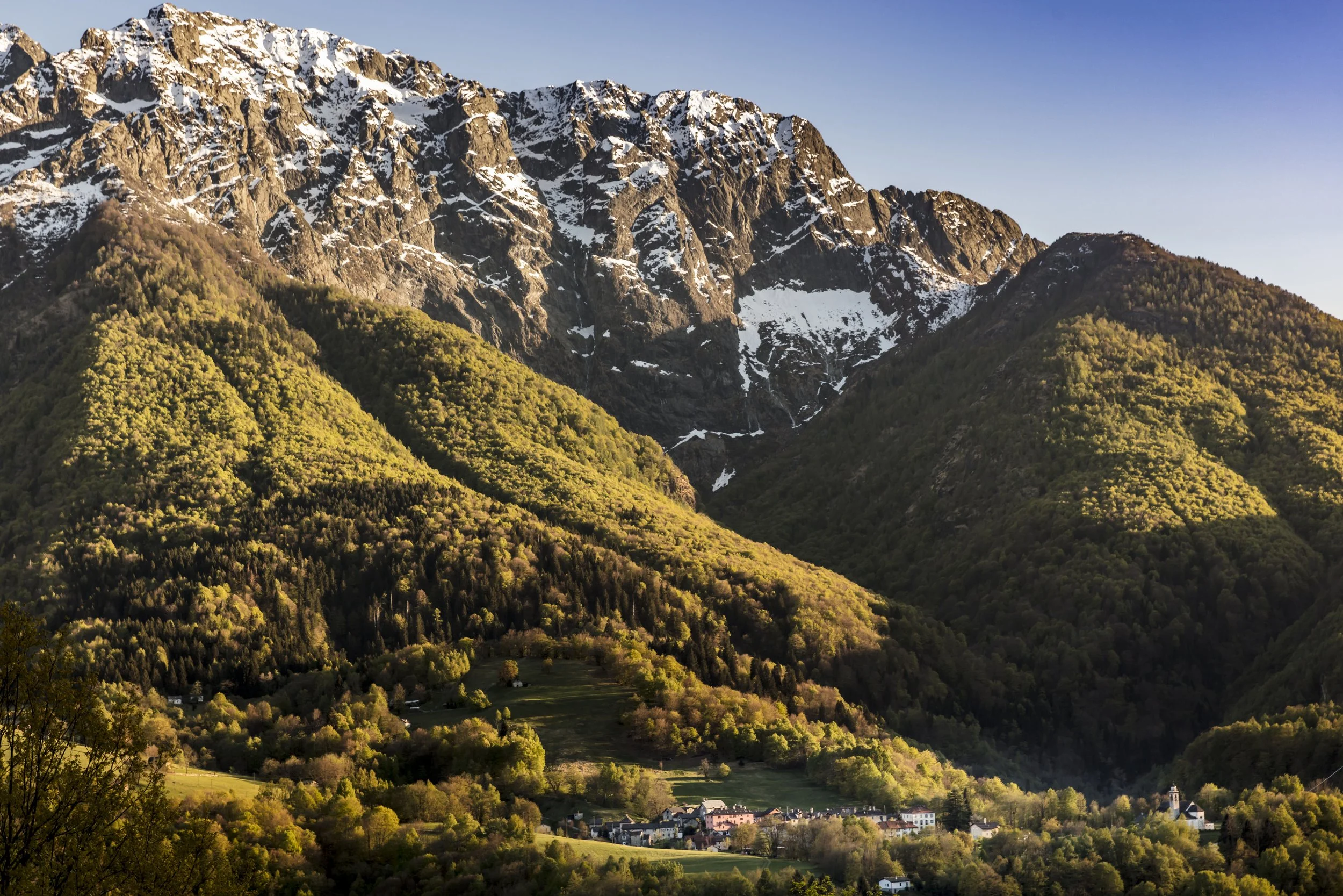 Paesaggio di montagne con cime innevate e colline verdi e boscosi, con un piccolo villaggio in basso.