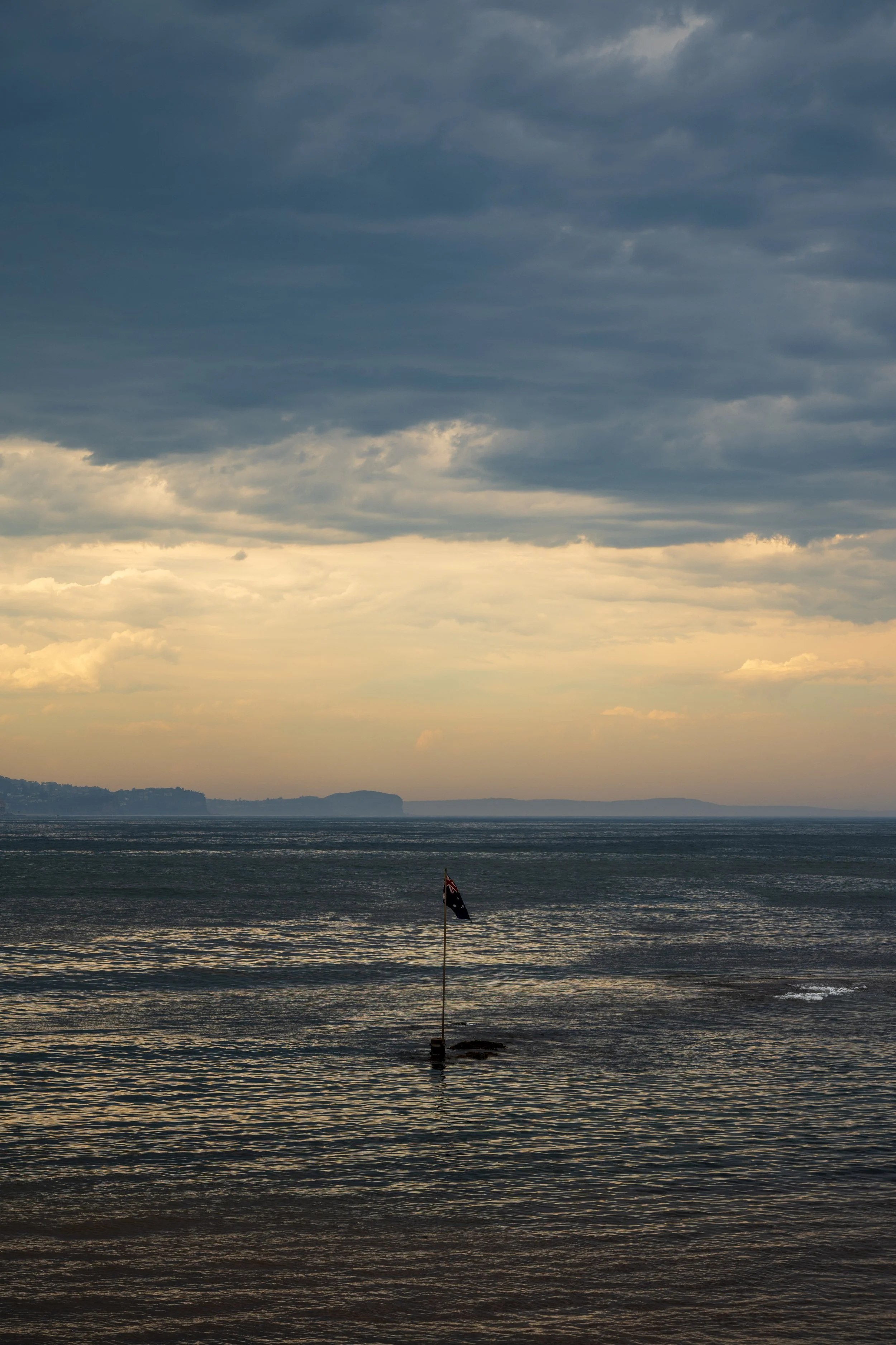 A coastal scene during cloudy weather, with a flagpole in the water and a small flag, and distant landforms on the horizon.