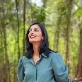 A woman with dark hair and a blue shirt standing outdoors surrounded by green trees.
