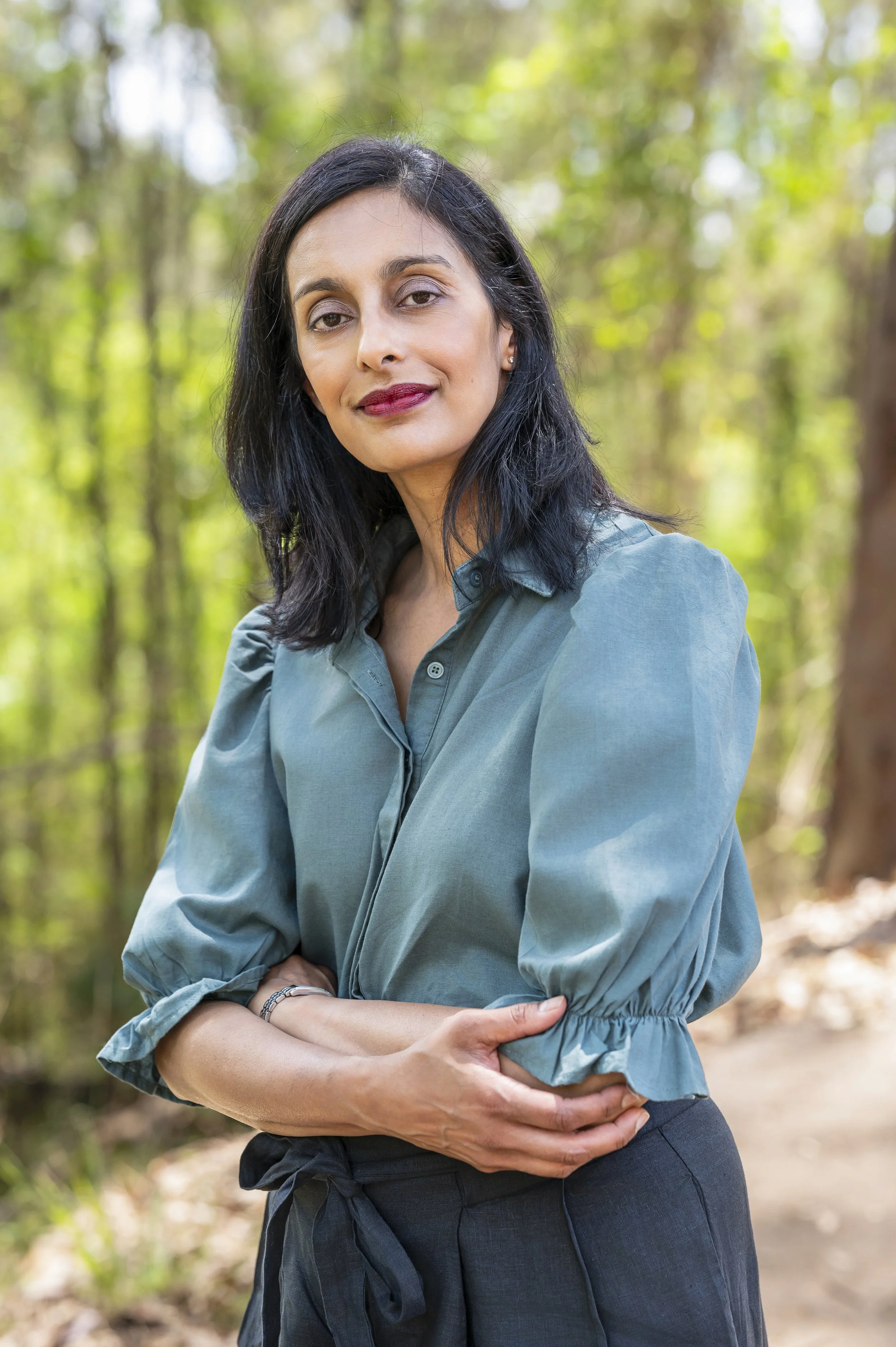 A woman with black hair and light makeup standing outdoors in a forest, wearing a teal blouse with puffed sleeves and black pants, looking at the camera with a slight smile.