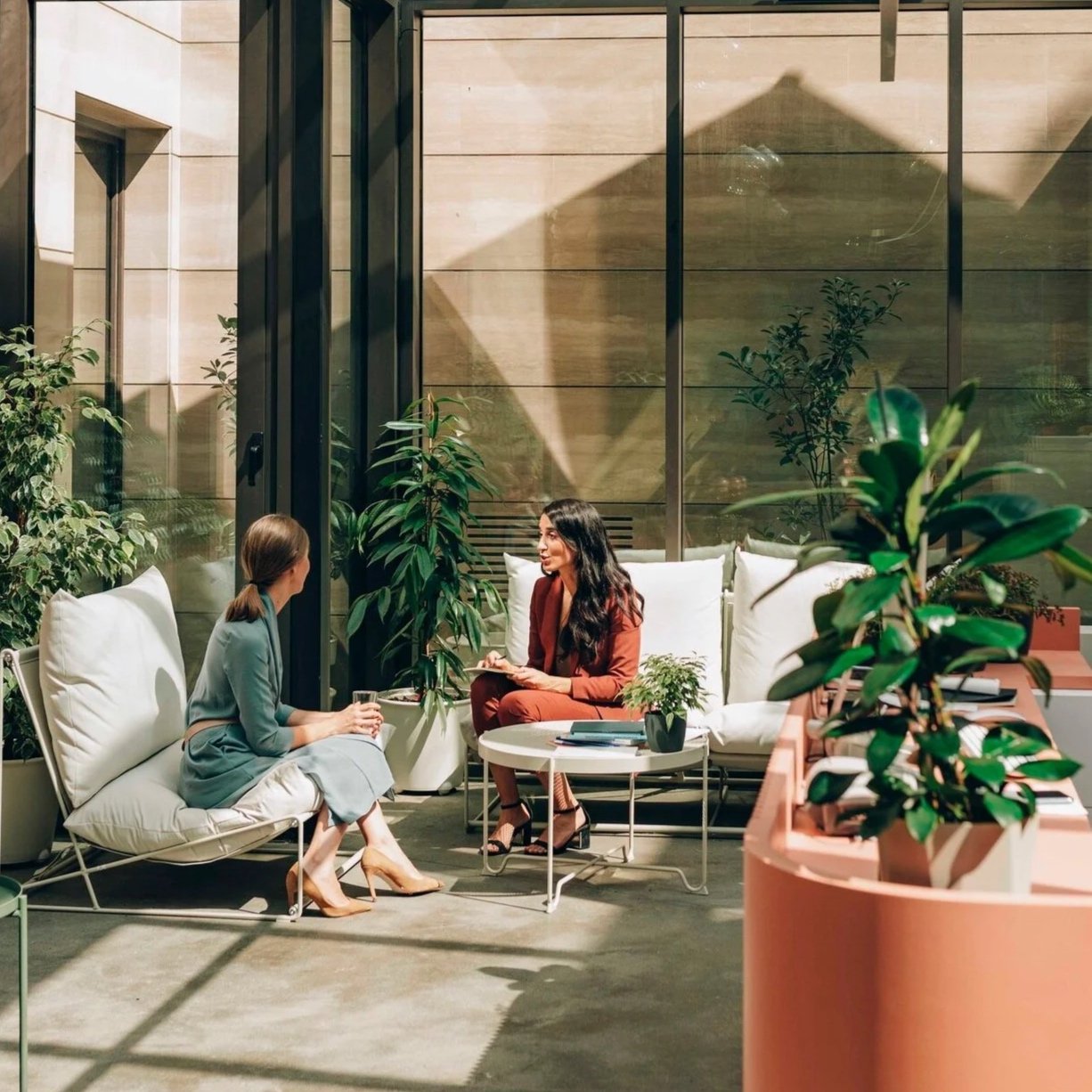 Two women seated on couches in a bright counselling office with high ceilings and plants, engaged in conversation, one holding a notepad and pen.