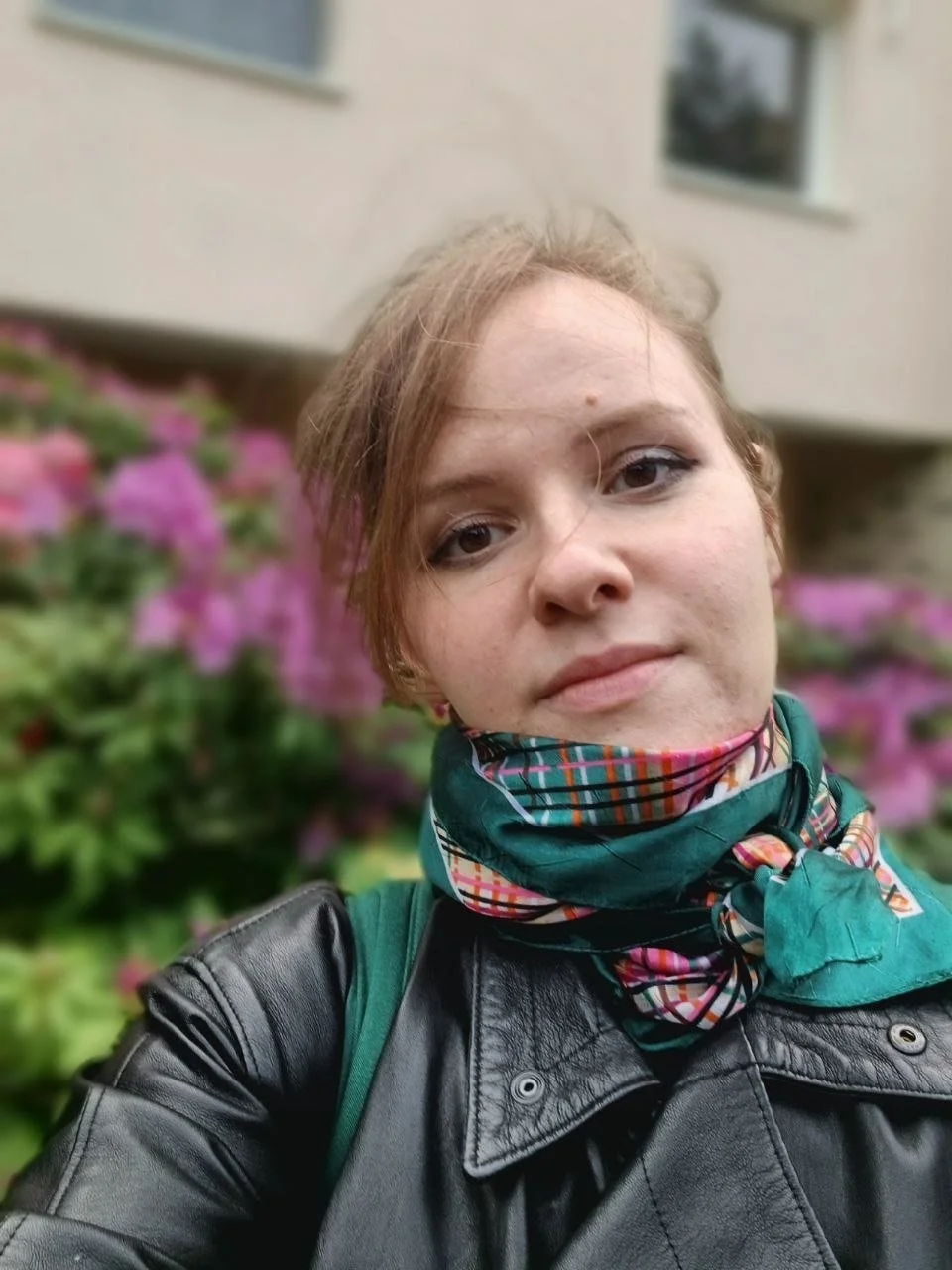 Eine junge Frau mit kurzen, roten Haaren trägt einen schwarzen Lederjacke und einen bunten Schal. Sie steht vor einem Hintergrund mit pinken Blumen und einem Gebäude.