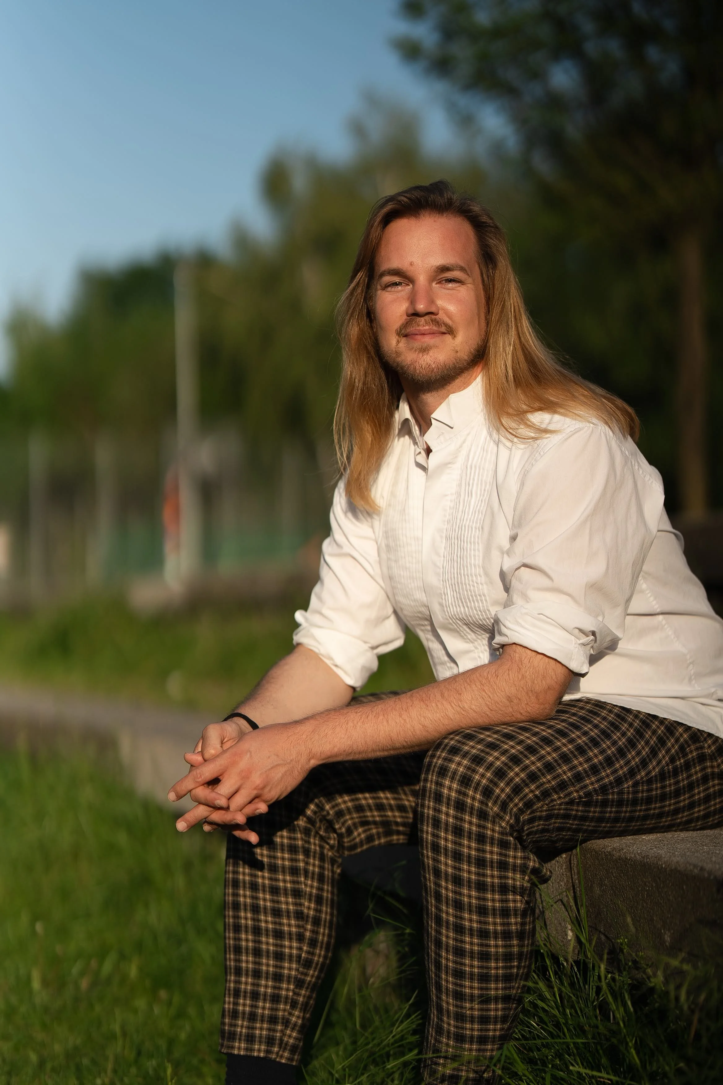 Ein Mann mit langen Haaren sitzt auf einem Steinhaufen im Freien, trägt ein weißes Hemd und karierte Hosen, und blickt entspannt in die Kamera, im Hintergrund Bäume und blauer Himmel.