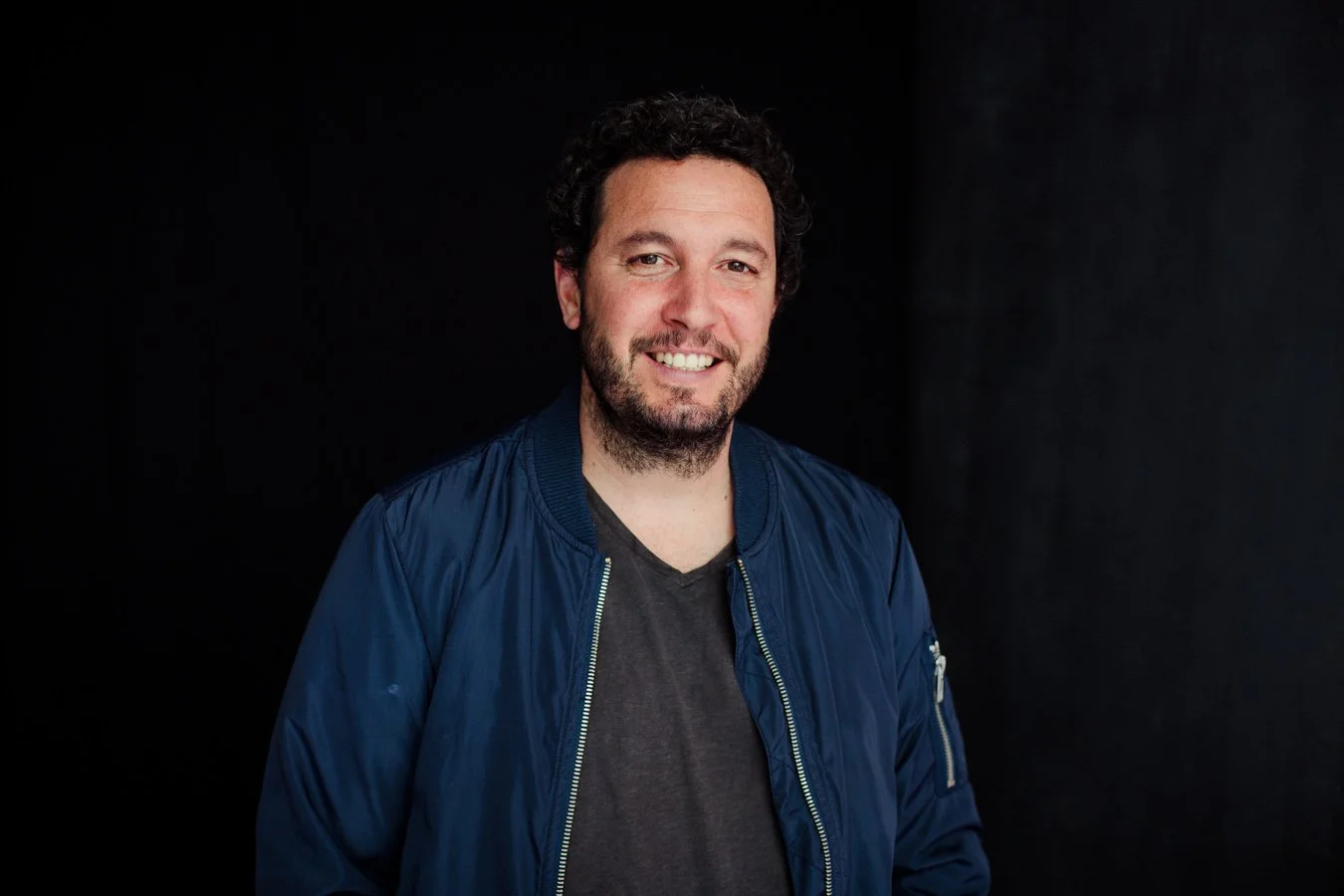 Ein lachender Mann mit dunklen, lockigen Haaren und Bart vor schwarzem Hintergrund, trägt eine blaue Jacke und ein dunkles T-Shirt.