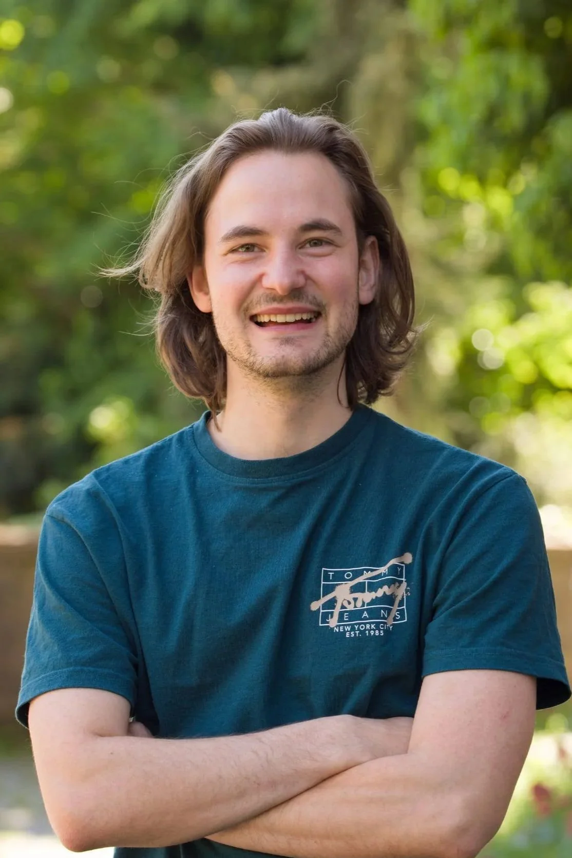 Lächelnder junger Mann mit langen braunen Haaren, trägt ein dunkelblaues T-Shirt. Er steht in einem grünen, bewaldeten Bereich.