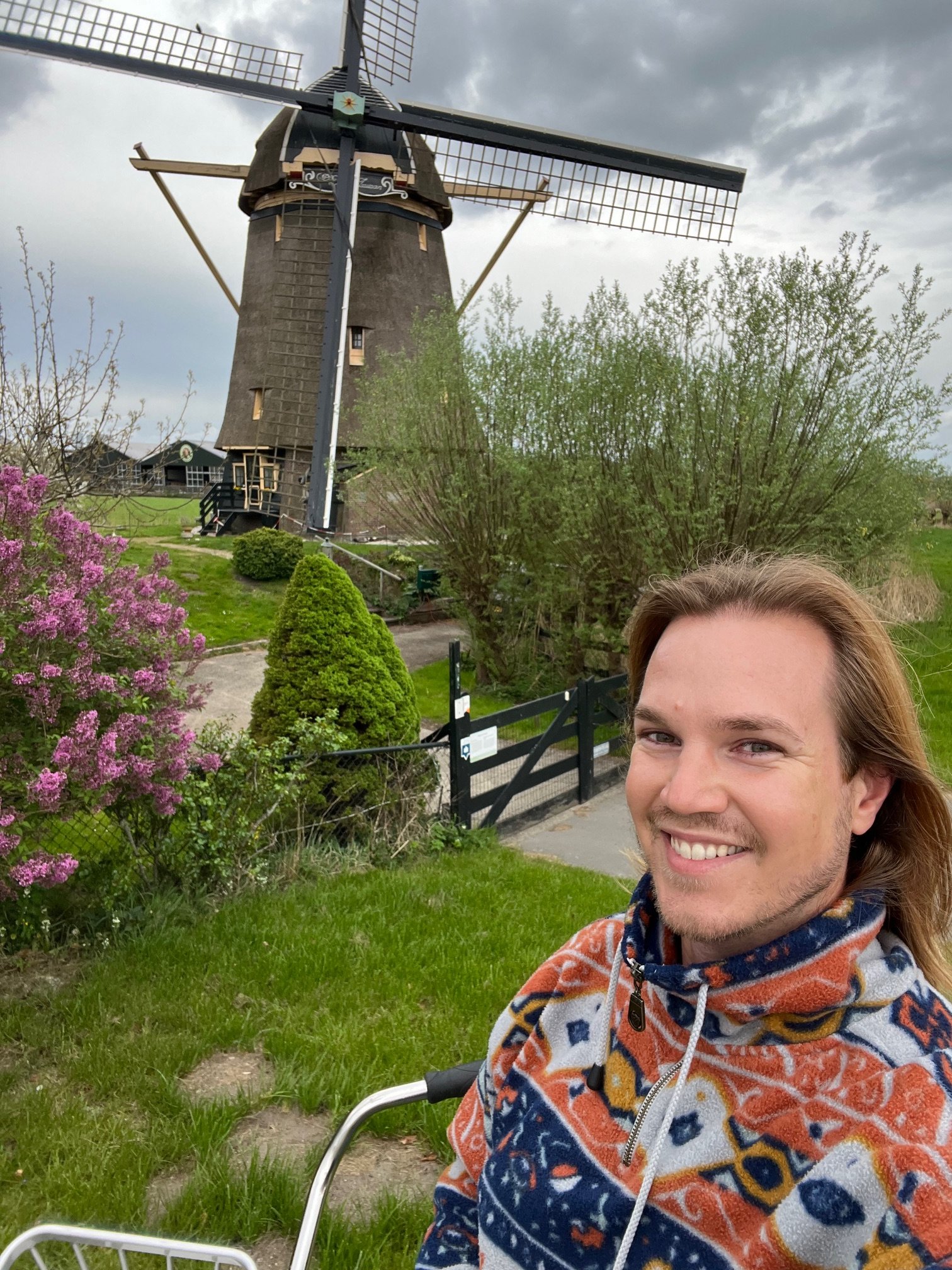 Ein lächelnder Mann im Vordergrund, im Hintergrund eine Windmühle auf einer grünen Wiese mit Büschen und Bäumen unter einem Wolkenhimmel.