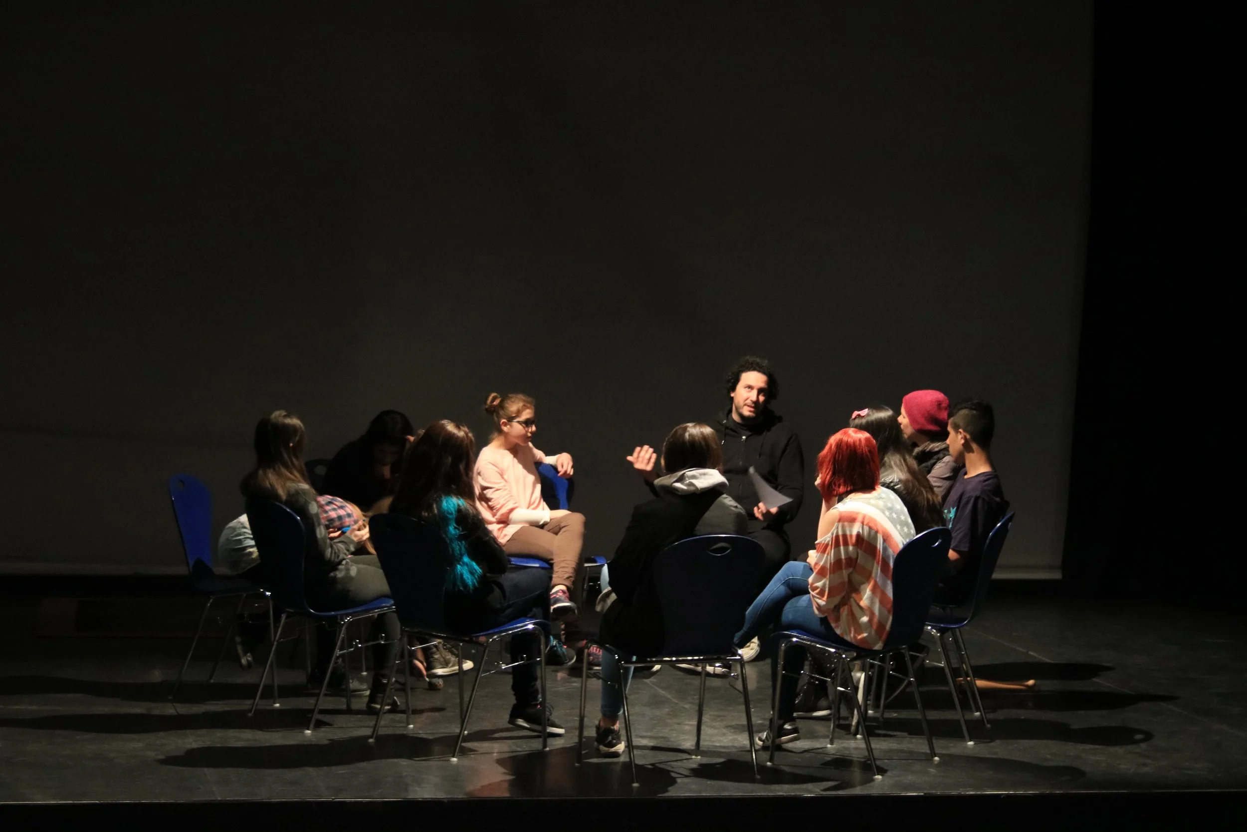 Eine Gruppe junger Menschen sitzt in einem Kreis auf Stühlen in einem dunklen Theaterraum. Ein Mann mit Locken spricht zu der Gruppe und erklärt etwas.